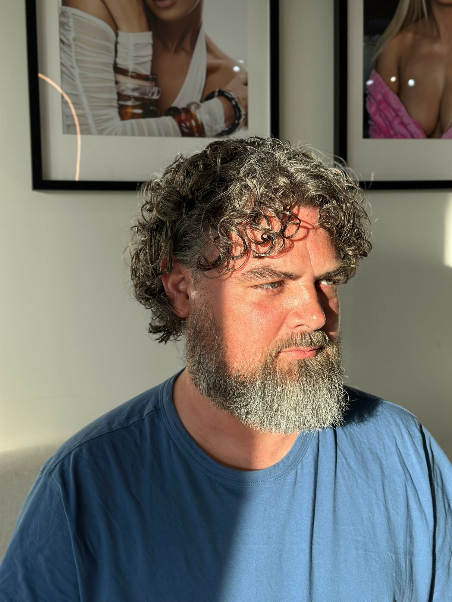 Man with curly hair and beard wearing a blue shirt, looking to the side, with framed photos on the wall behind him.