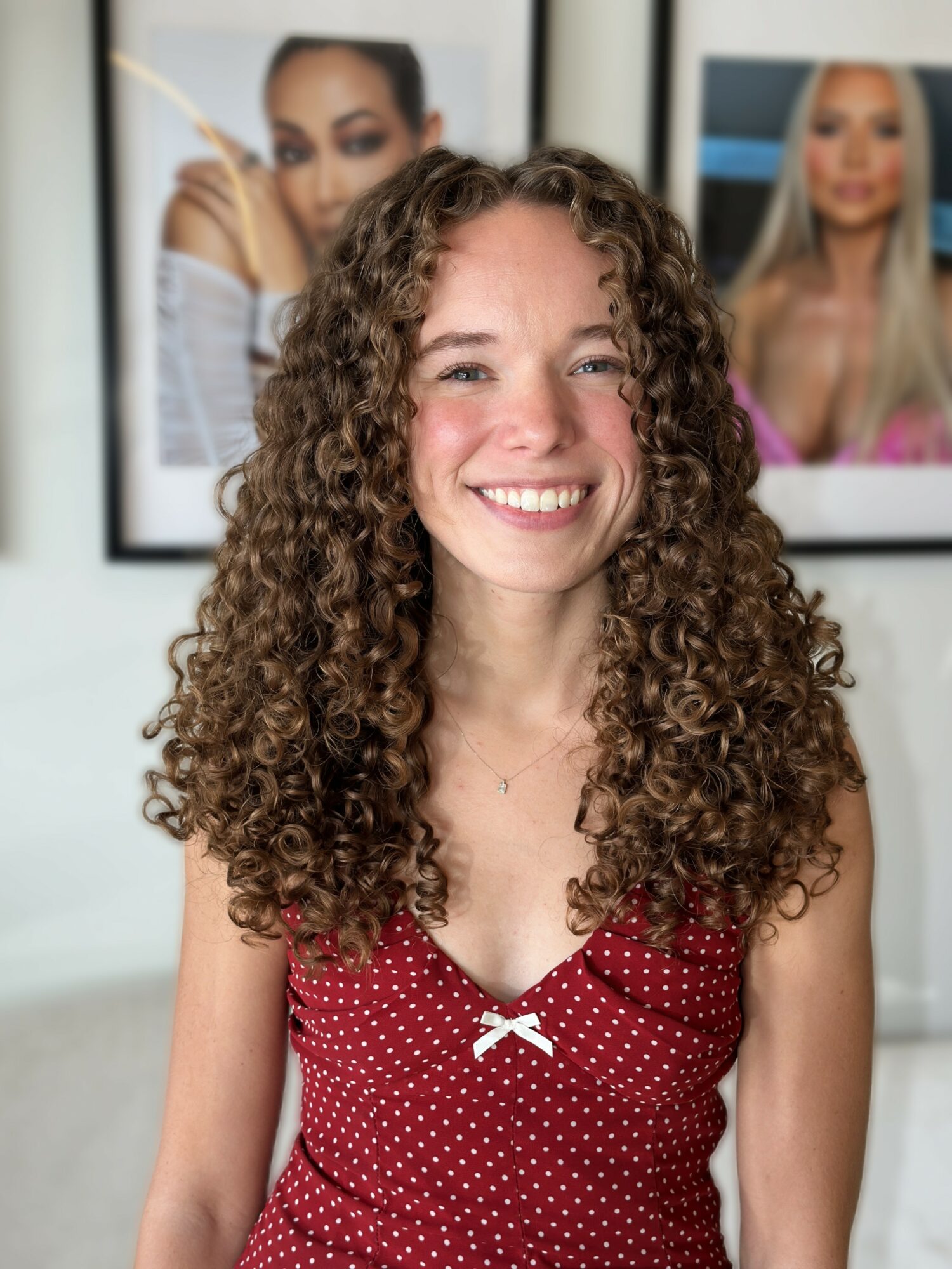 Young woman with curly hair smiling, wearing a red polka dot top, with framed photos of women in the background.