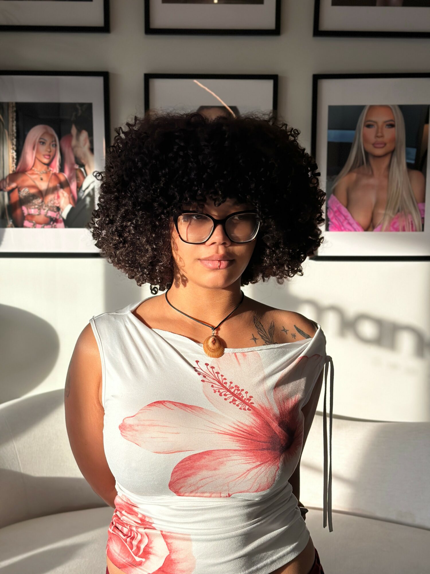 Woman with curly hair and glasses, wearing a white top with a pink flower design, in an art gallery.
