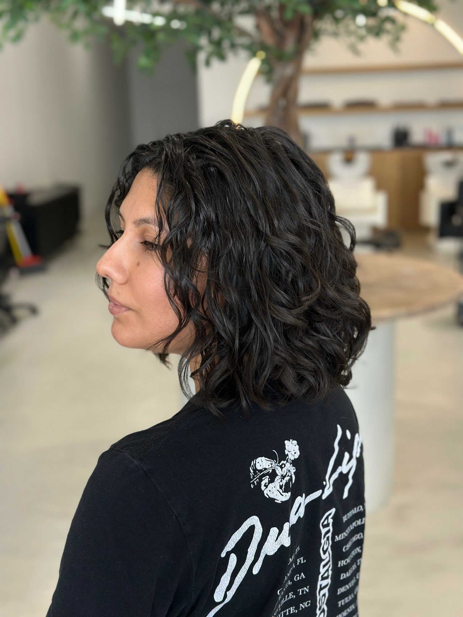 Side profile of a woman with curly dark hair in an indoor setting, background includes a tree and furniture.