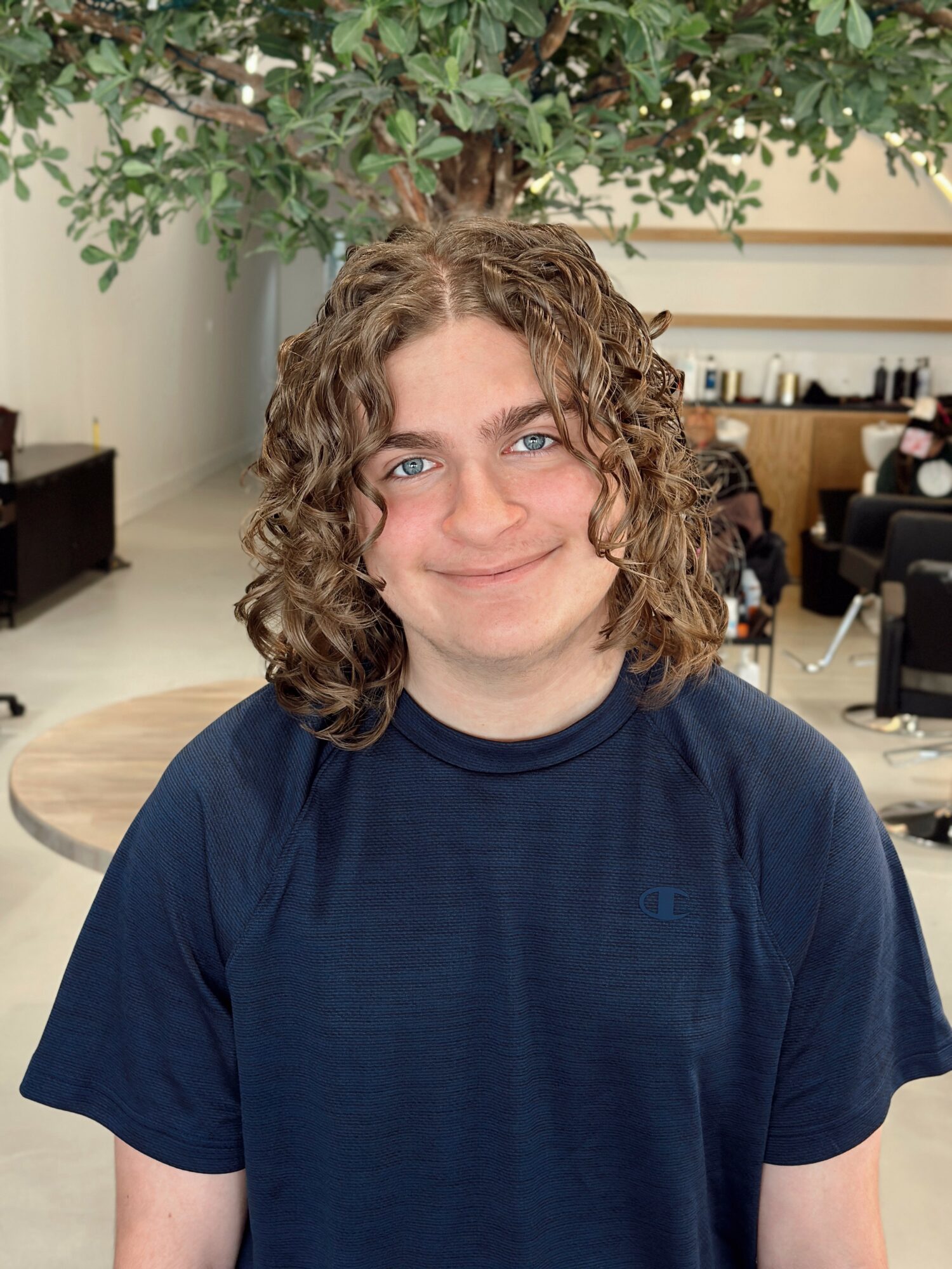 Young person with curly hair smiling, wearing a dark blue shirt, indoors with plants and furniture in background.