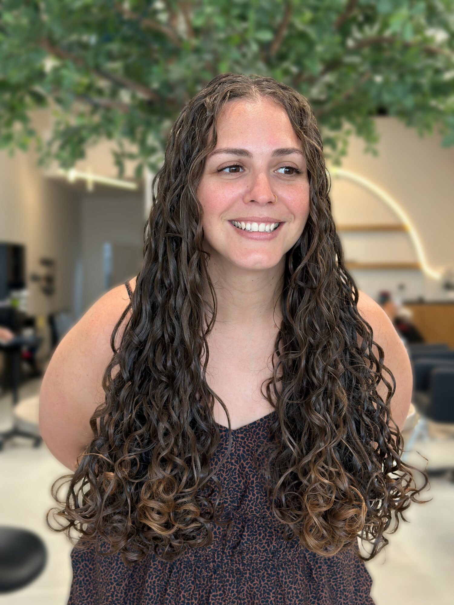 Young woman with long, curly hair smiling indoors, background with trees and modern architecture.