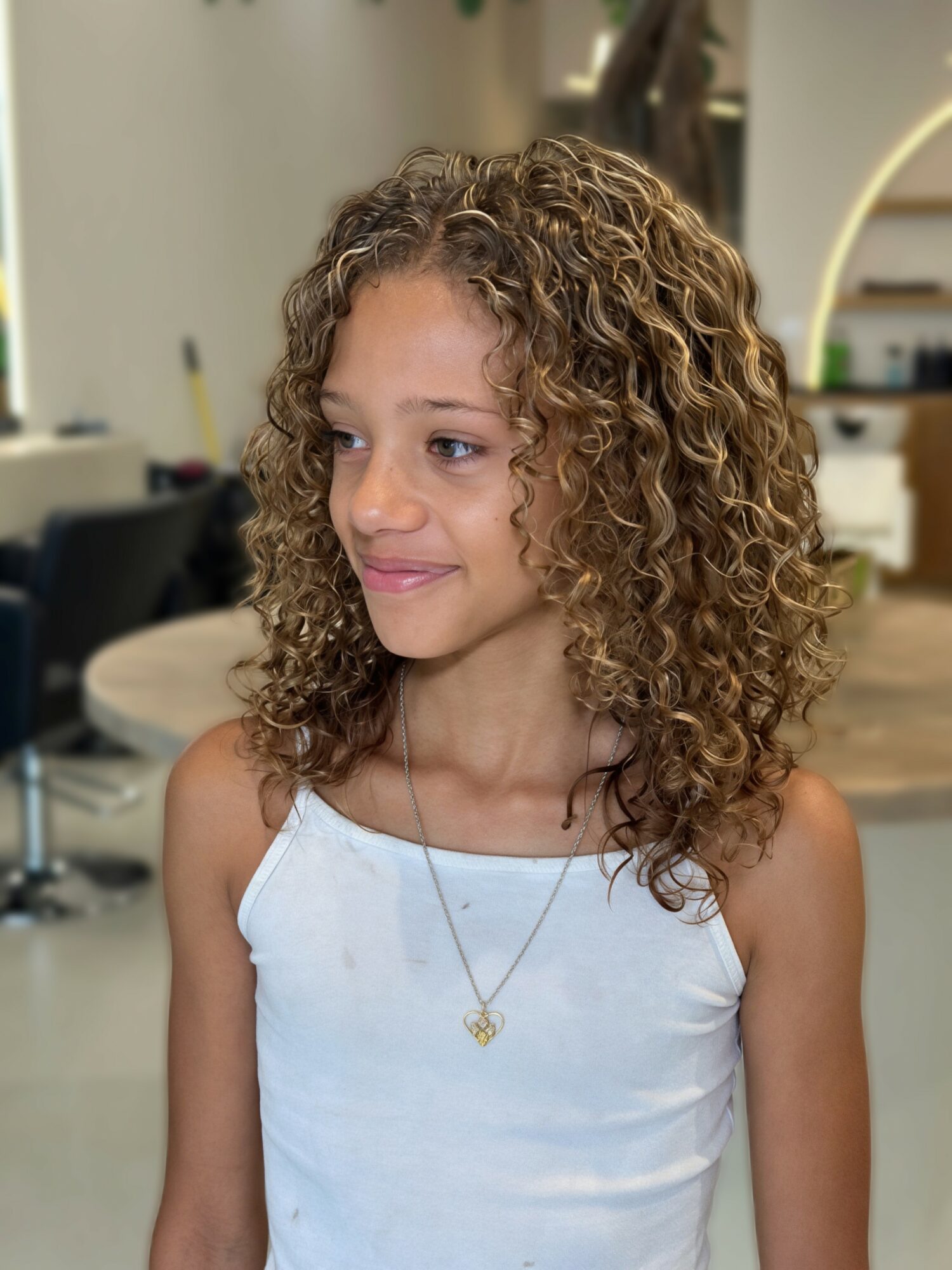 Young girl with curly hair wearing a white sleeveless top and a necklace, smiling softly indoors.