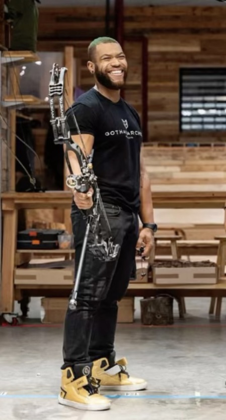 Person with a prosthetic arm holding a bow, smiling, in a wooden indoor space.