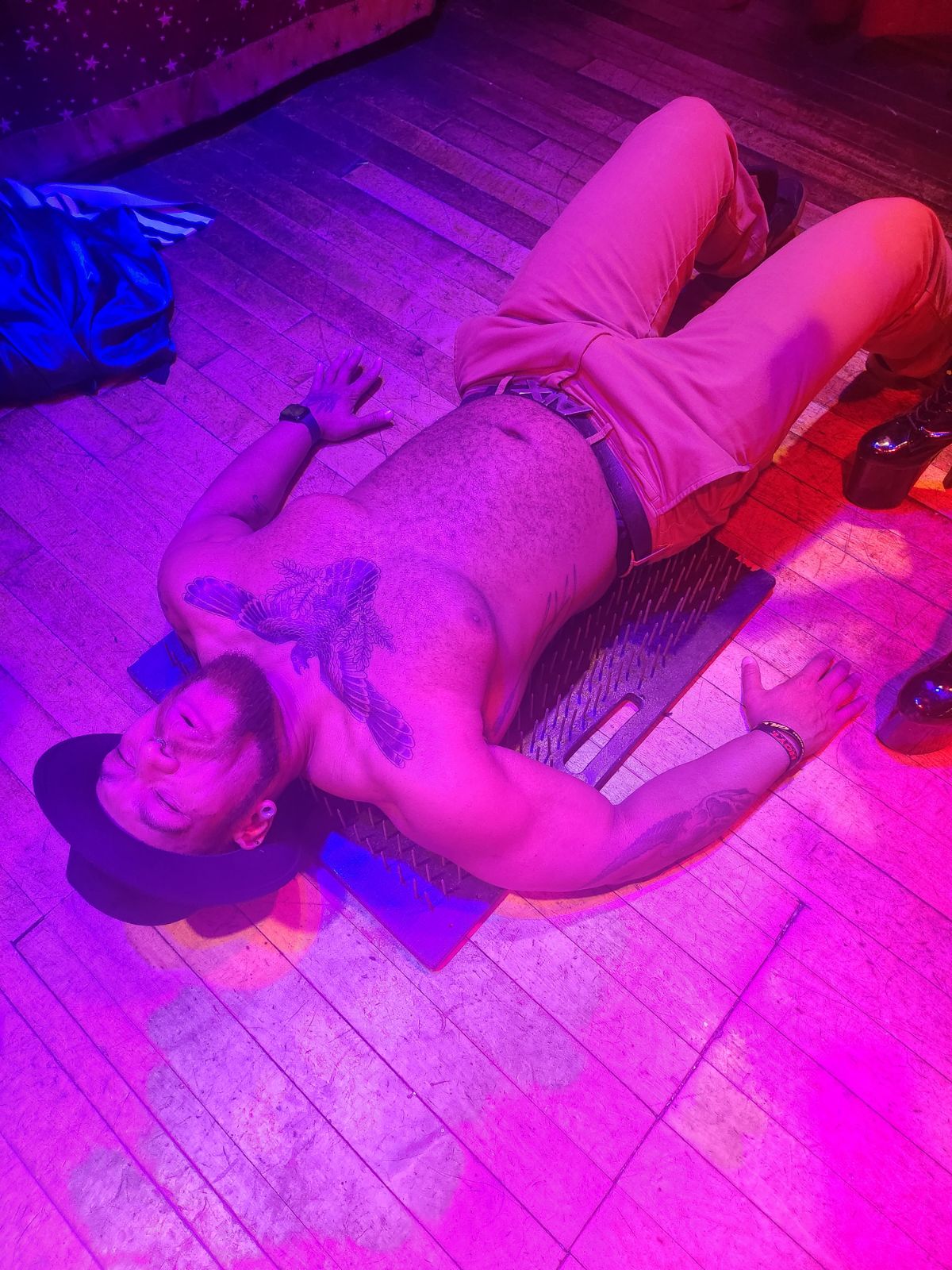 Person lying on a mat on the floor, shirtless, wearing shorts, with a hat, in a room with wooden flooring and purple lighting.