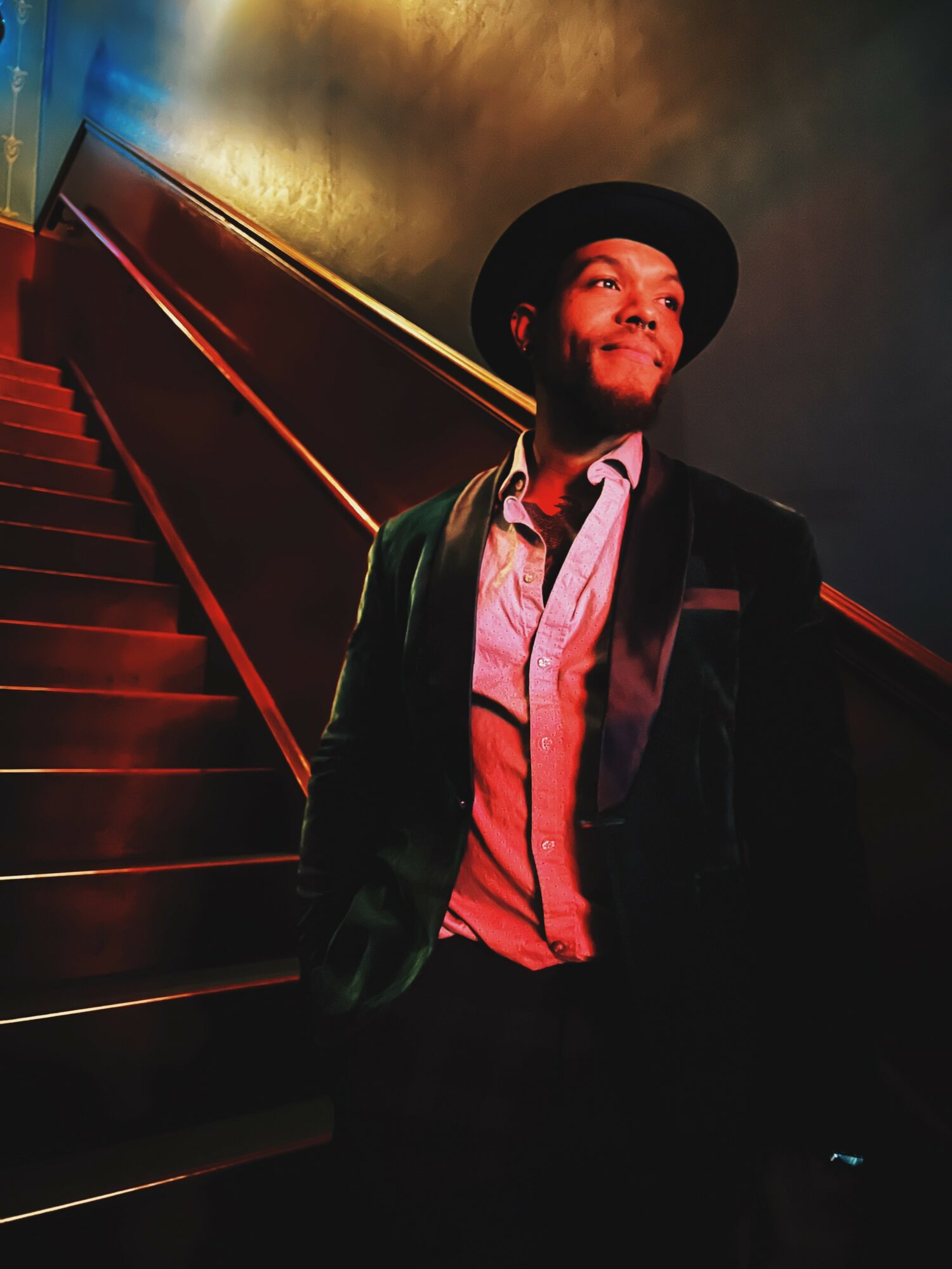 Person wearing a hat and jacket standing on stairs in dim lighting, looking to the side with a serious expression.
