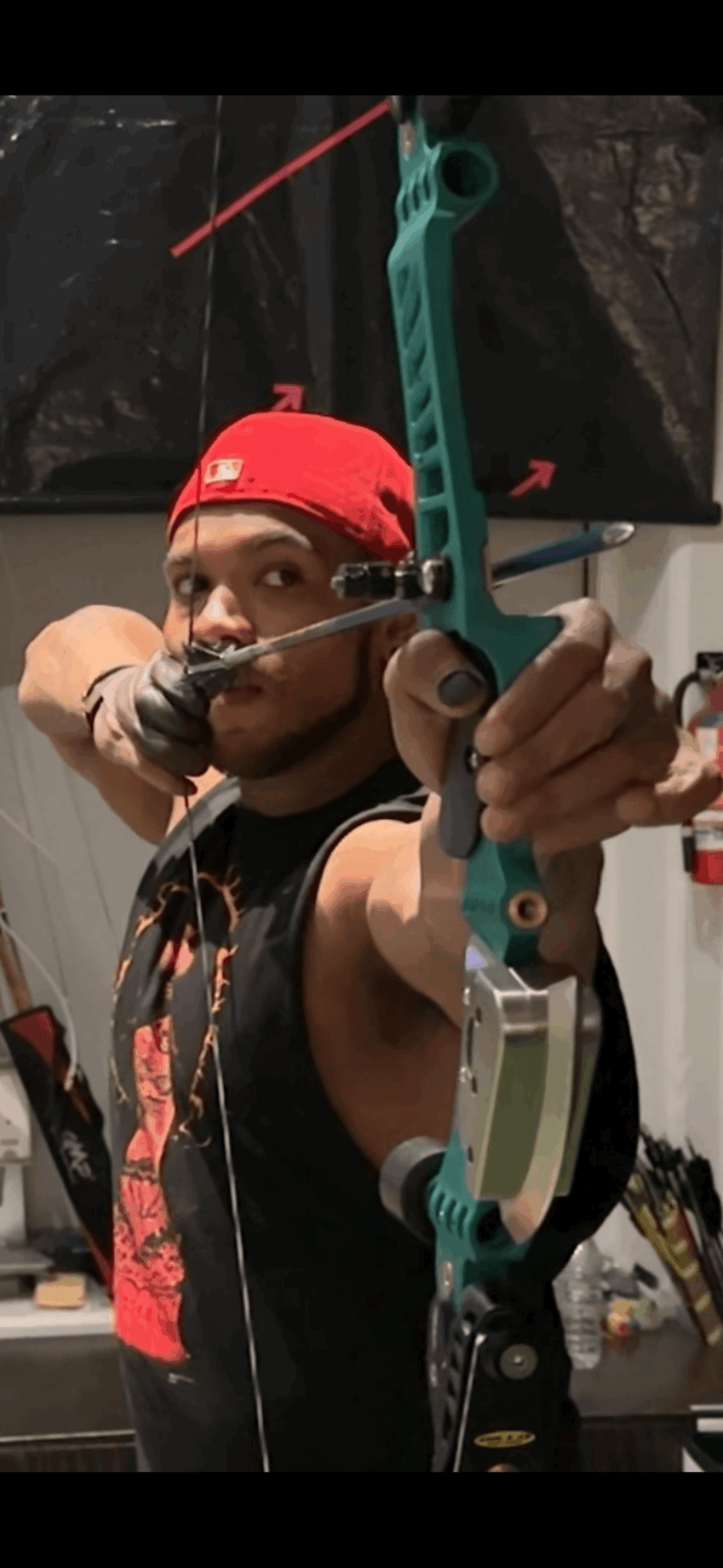 Person wearing a red cap and black sleeveless shirt aiming a toy bow and arrow indoors.