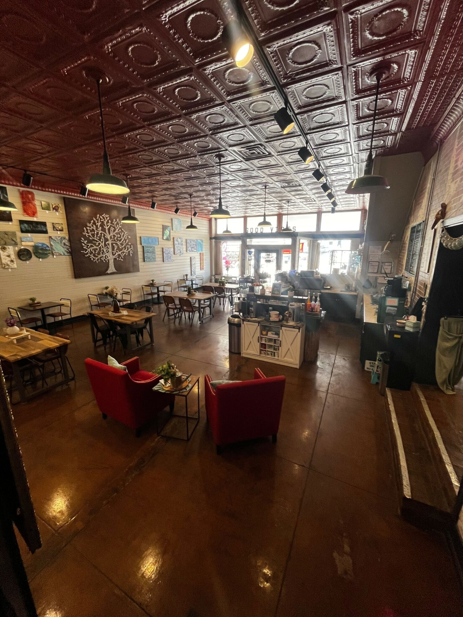 Interior of a cafe or restaurant with a patterned ceiling, wooden floor, tables, chairs, and a bar area near large windows.