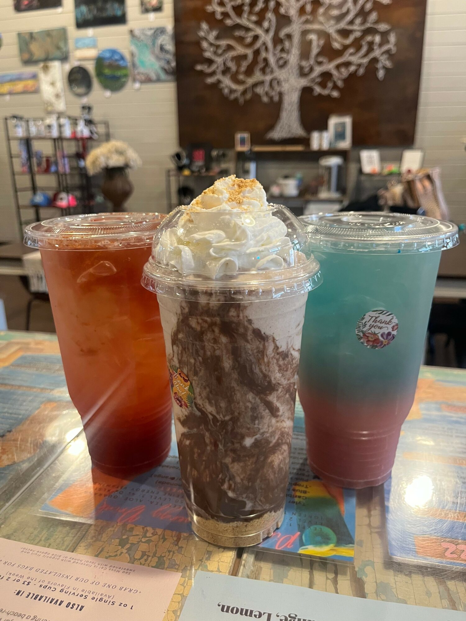 Three cold beverages in plastic cups with whipped cream, chocolate, and ice, on a table in a cafe or restaurant.