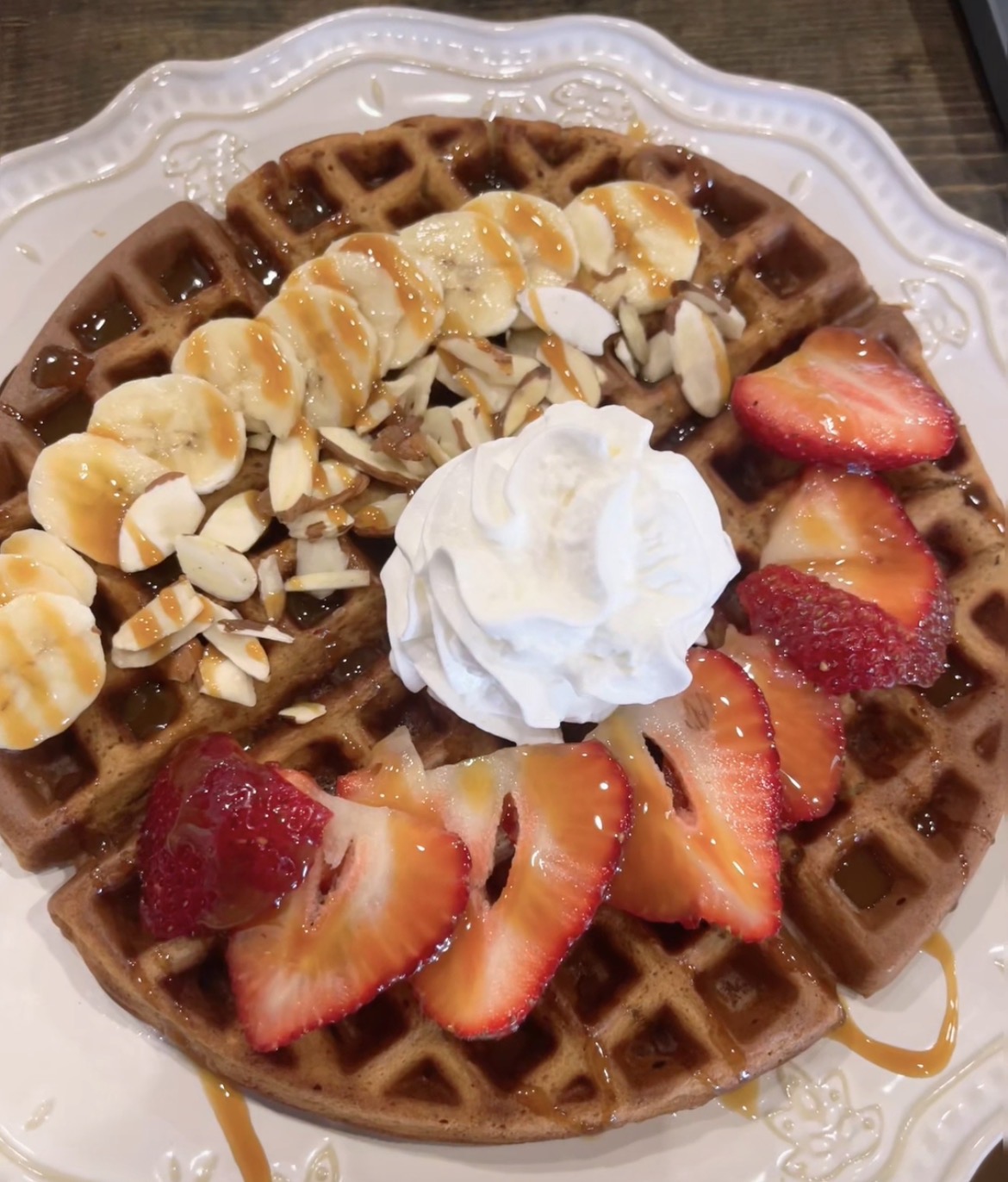 Waffle topped with sliced bananas, strawberries, whipped cream, and almond slices, drizzled with syrup on a decorative plate.