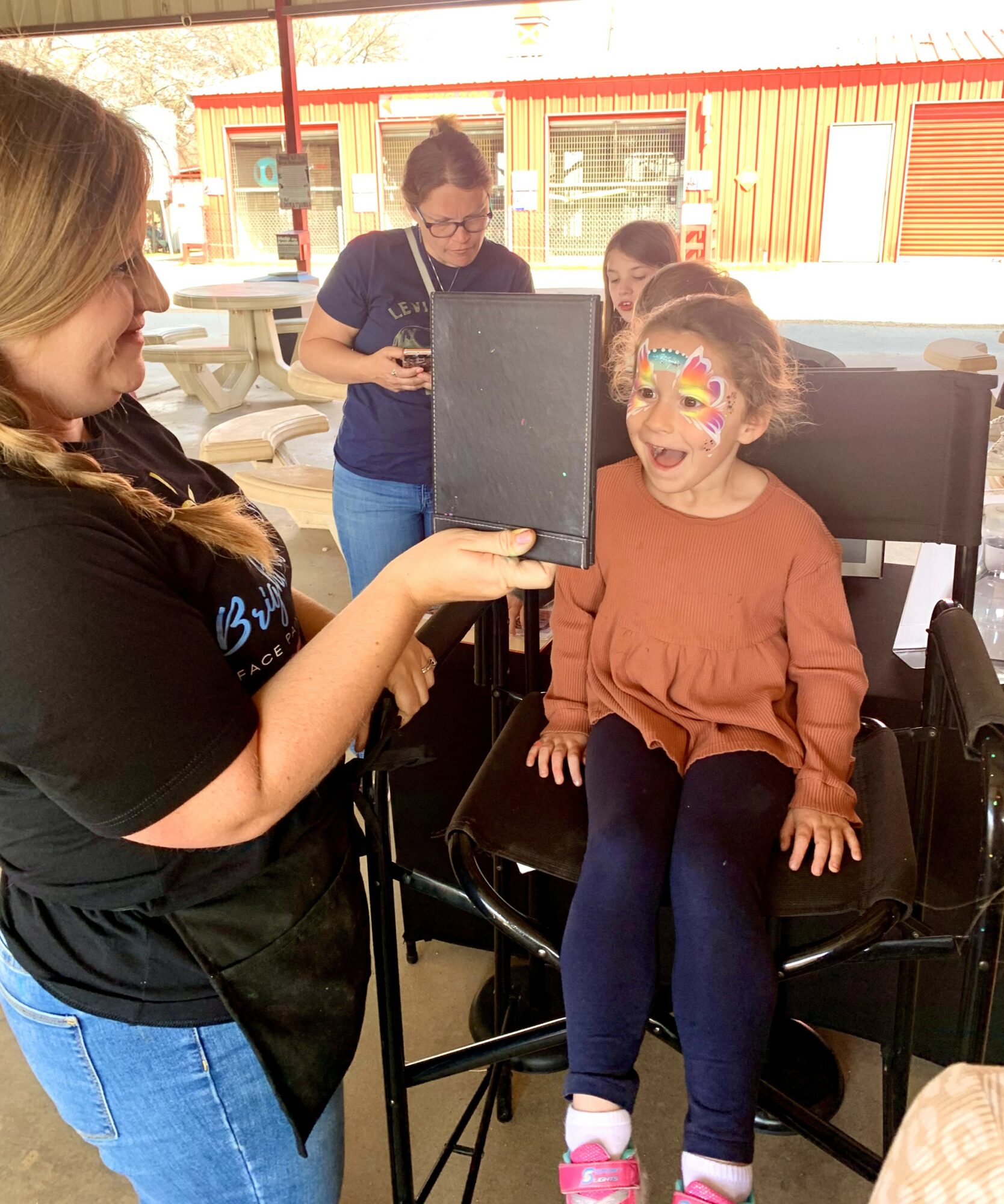 Girl in wheelchair with face paint, smiling, while woman takes her photo outdoors with a tablet, group in background, sunny day.