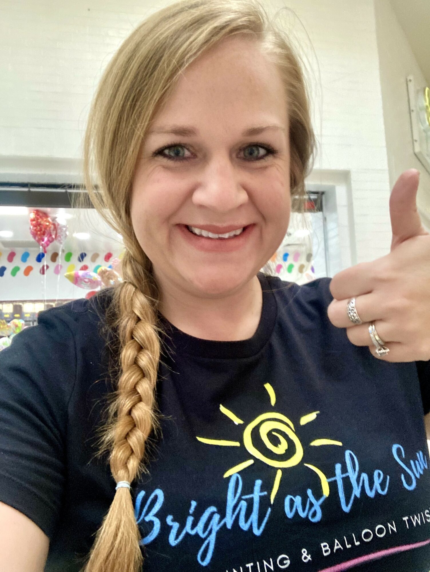 Smiling woman with long braided hair giving a thumbs-up, wearing a dark T-shirt with colorful text, indoors.