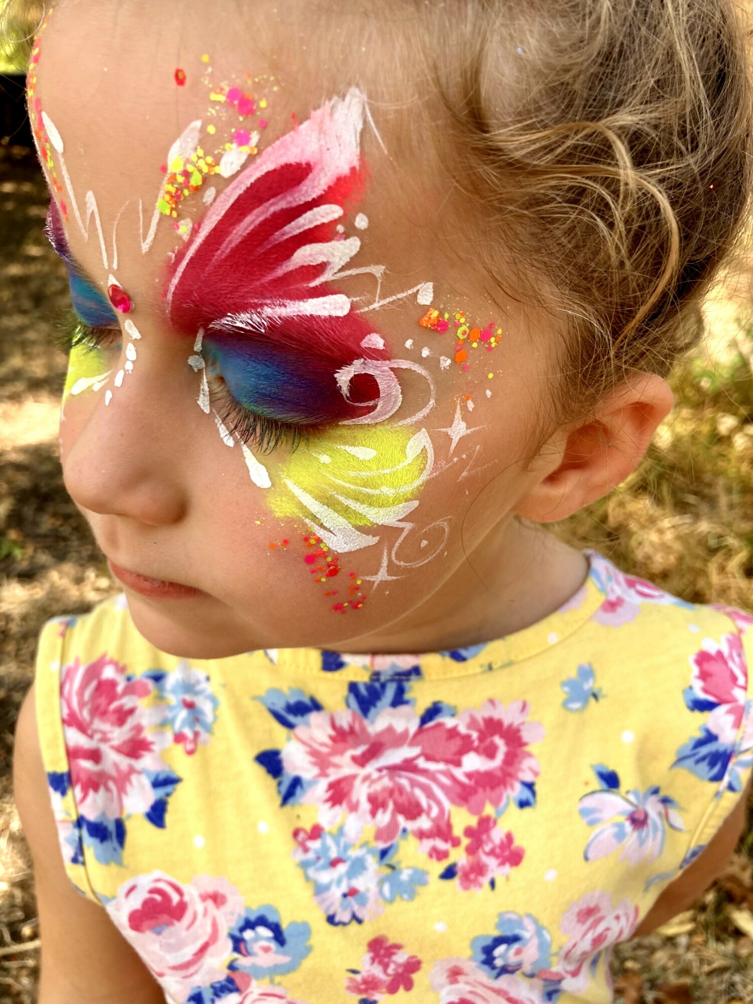 Child with colorful butterfly face paint, wearing a yellow floral dress, outdoors with trees in background.