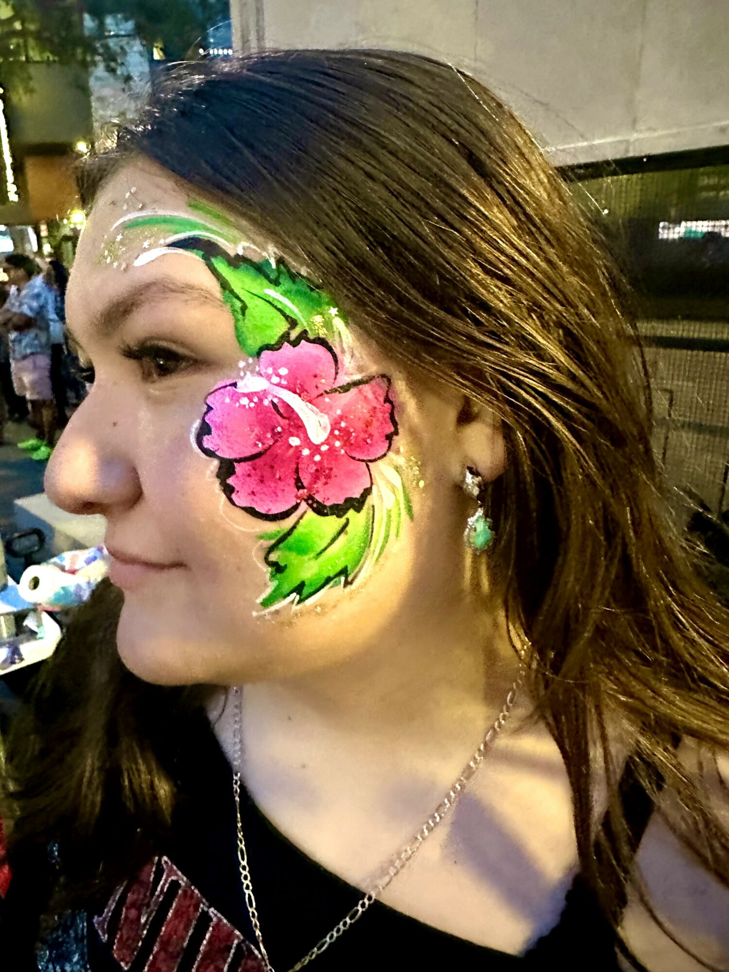 Side profile of a woman with face paint of pink flower and green leaves on her cheek, outdoor setting at night.