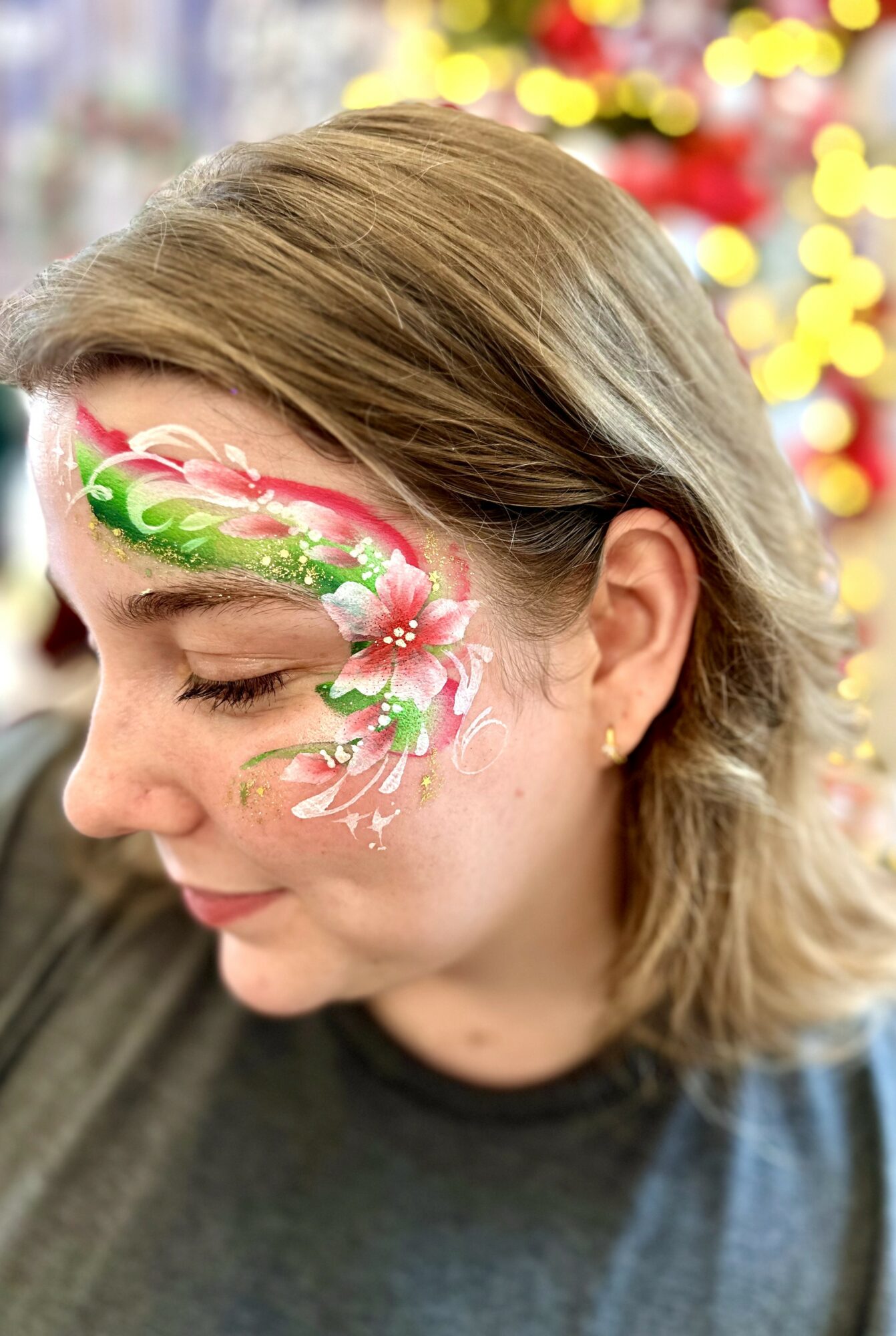 Woman with Christmas-themed face paint featuring a pink and green design on her cheek, smiling with eyes closed.