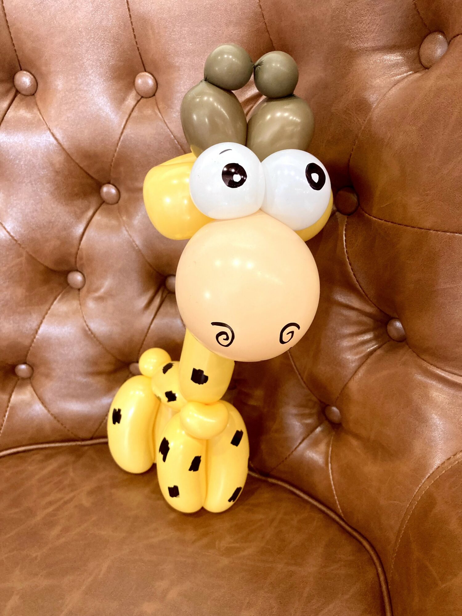 Balloon sculpture resembling a giraffe with a large beige head, yellow body, and green ears, on a brown tufted surface.
