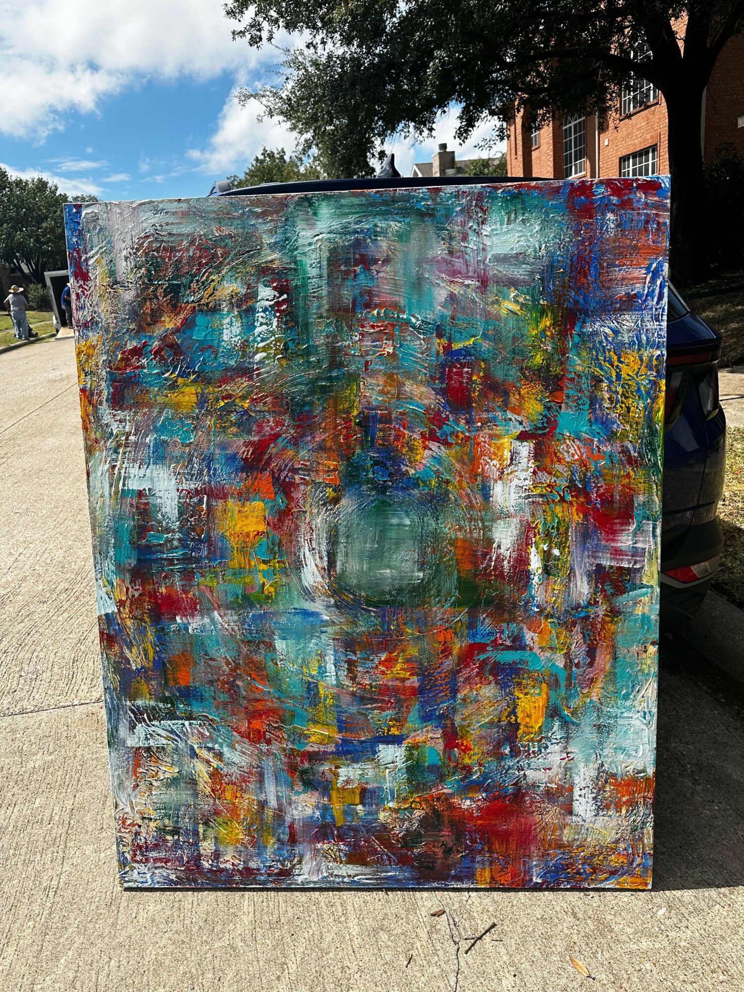 Colorful abstract painting with various brushstrokes and textures, outdoors on a sidewalk, with trees and buildings in the background.