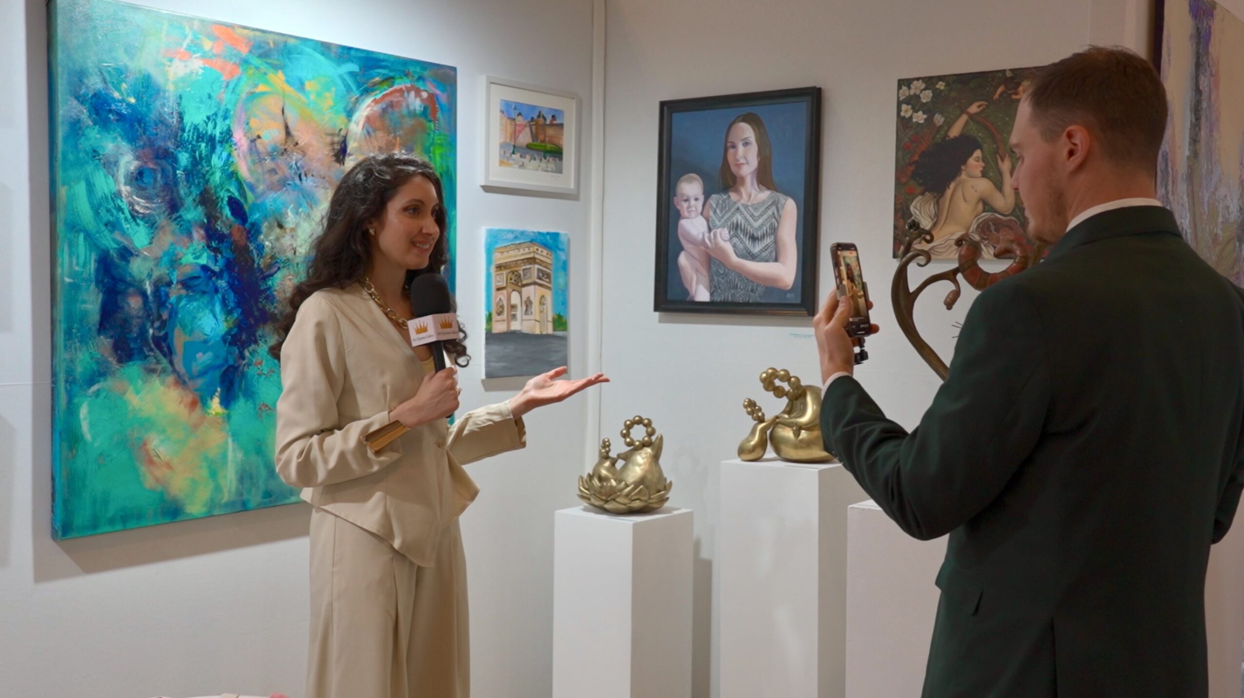 Woman being interviewed at an art gallery with paintings and sculptures displayed behind her.