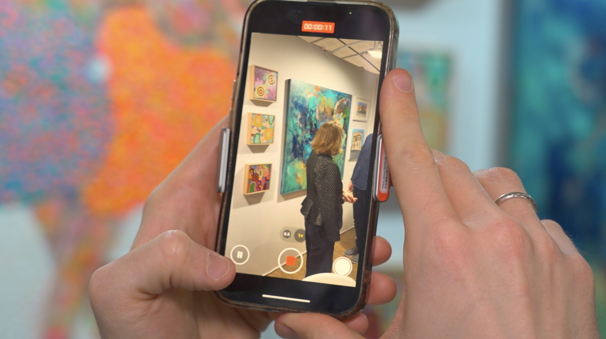 Person taking a photo of artwork in an art gallery with a smartphone, colorful paintings in background.