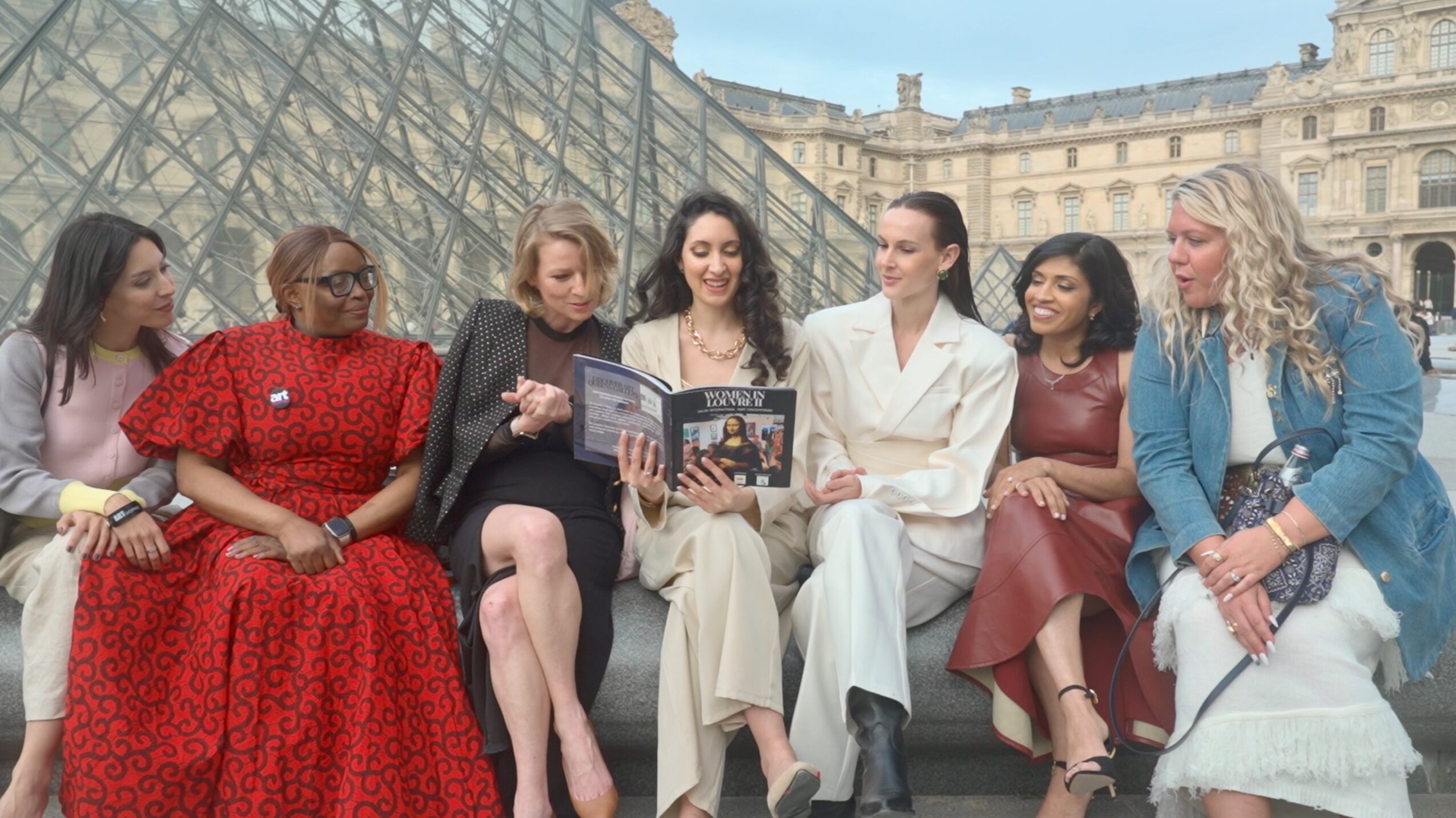 Seven women sitting on a bench outdoors, looking at a magazine, with a building and glass structure in the background.