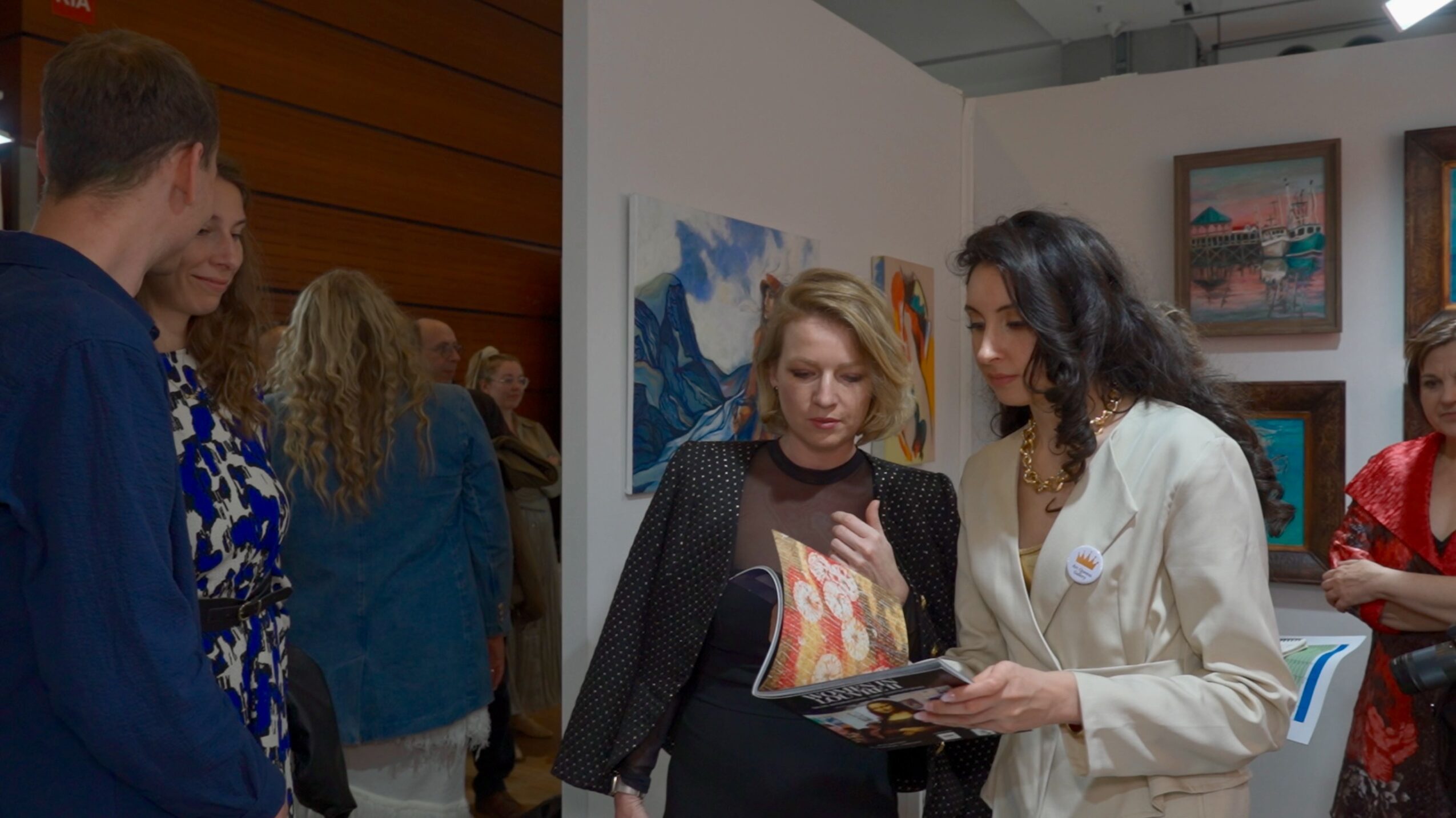Two women look at a book or catalog while others observe in an art gallery or exhibition space.
