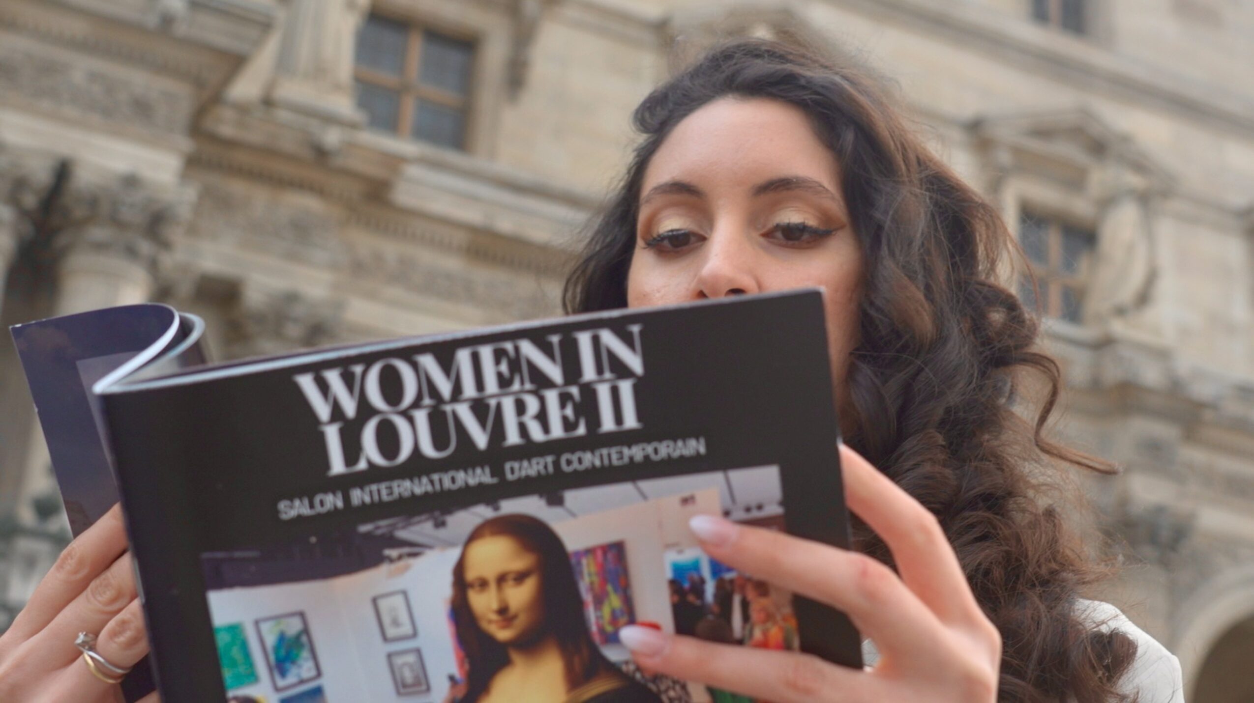 Woman with curly hair reading a book titled 'Women in Louvre II' outdoors, with historic building in background.