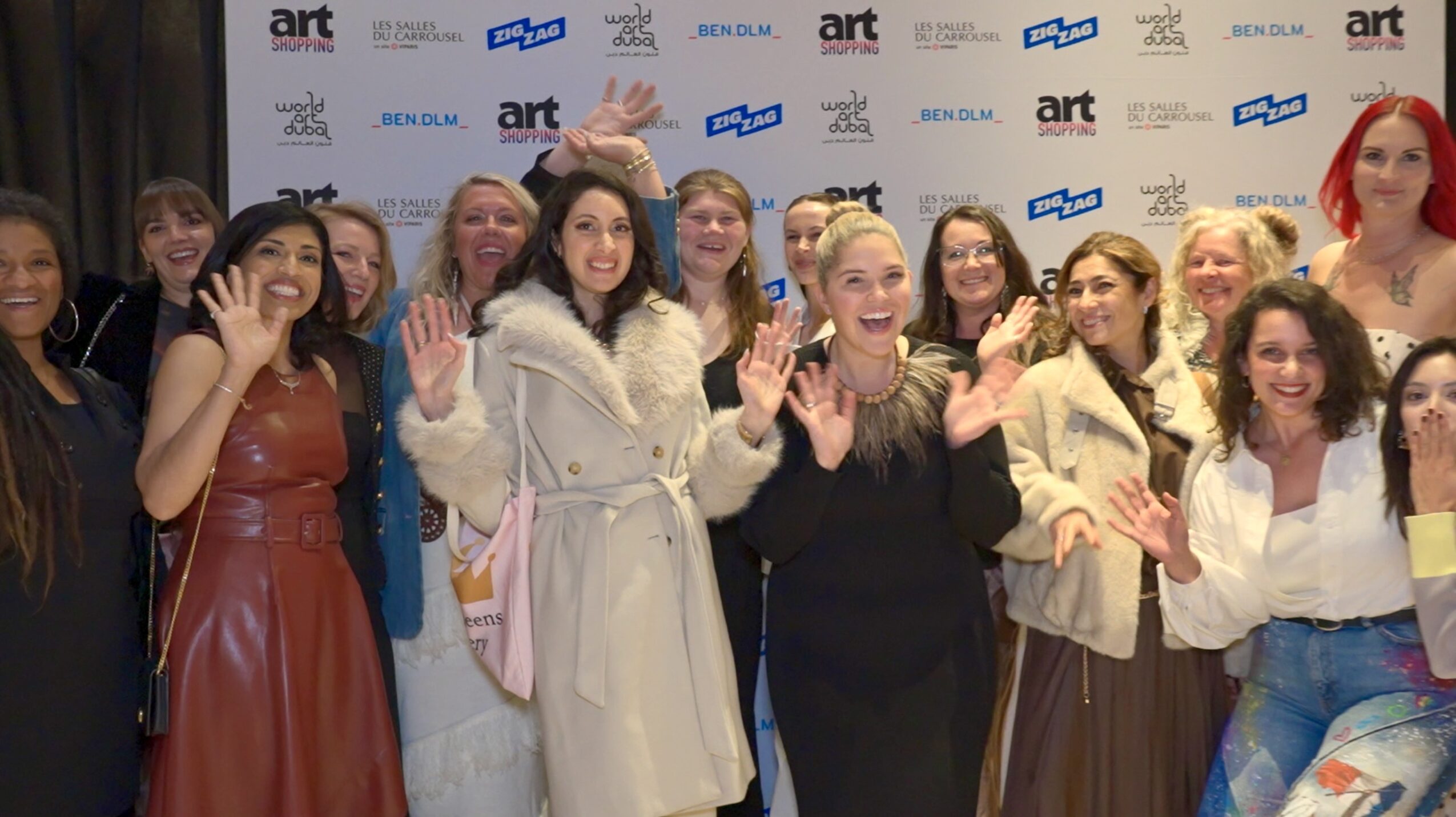 Group of women smiling and waving at an event with a backdrop of logos and text.