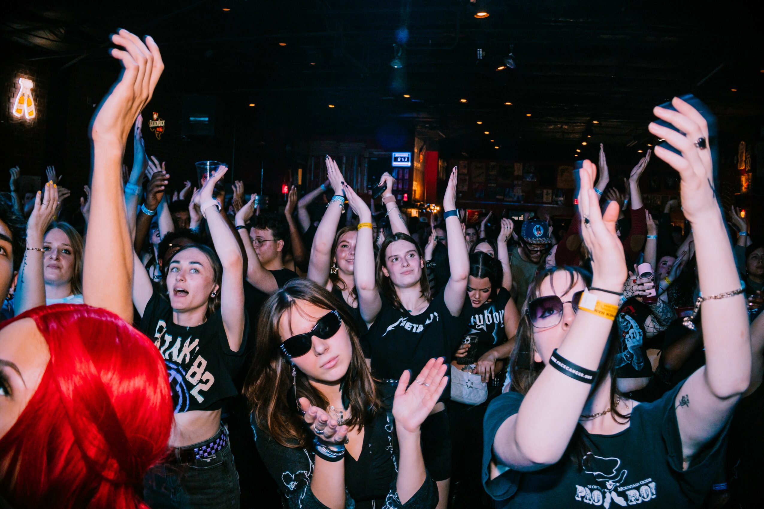 Crowd of people at a concert or event, many with hands raised, some wearing sunglasses, in a dark indoor setting.