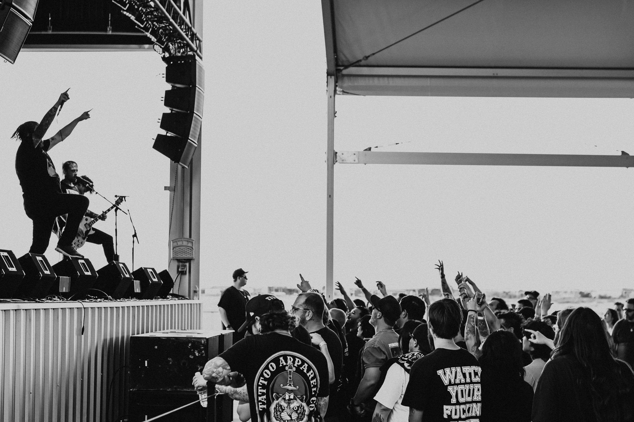 People at an outdoor event with a stage, some raising hands, and a large open sky in the background.