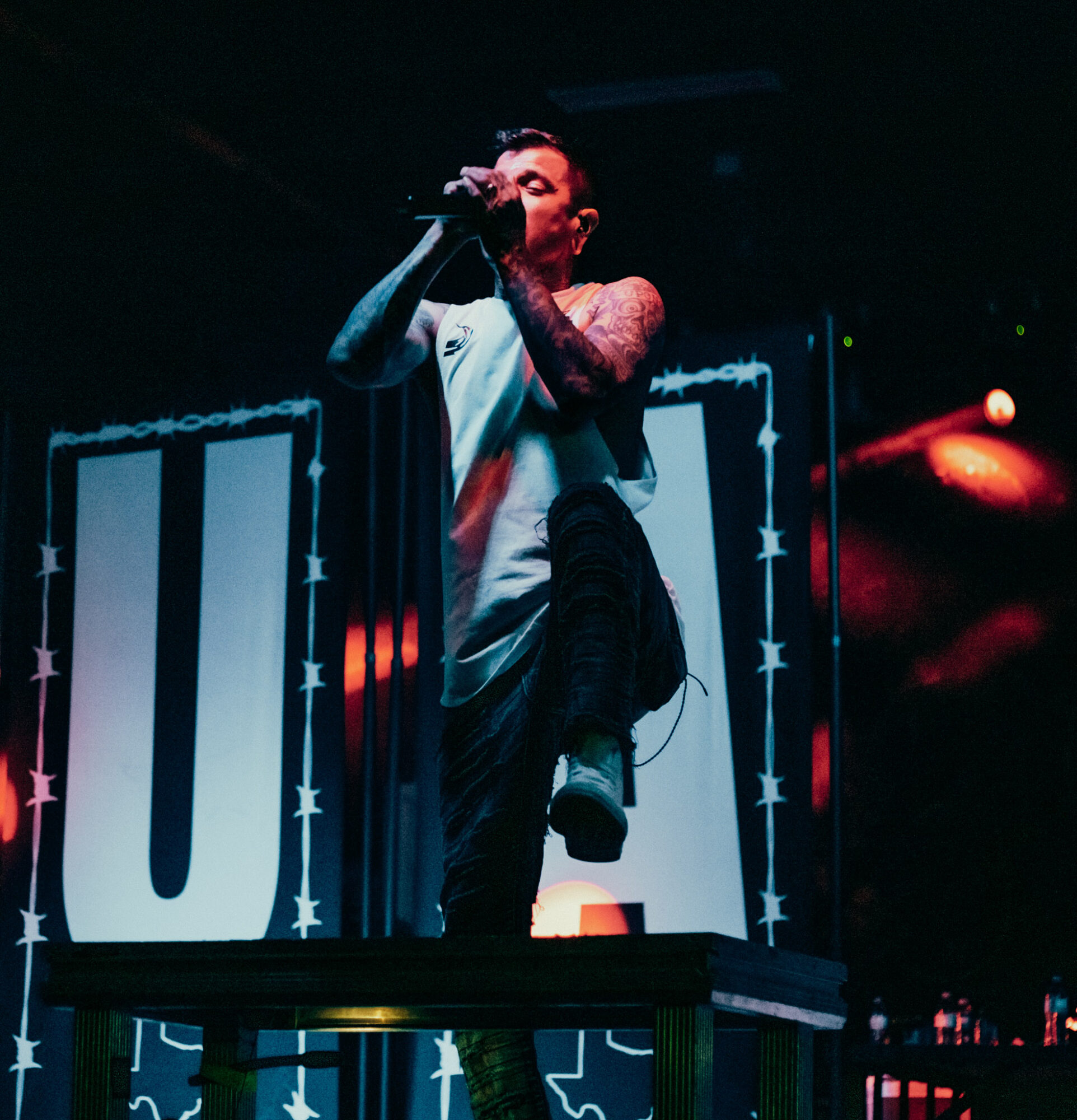 Male performer singing into microphone on stage with illuminated background, standing on a table, tattoos visible on arms.