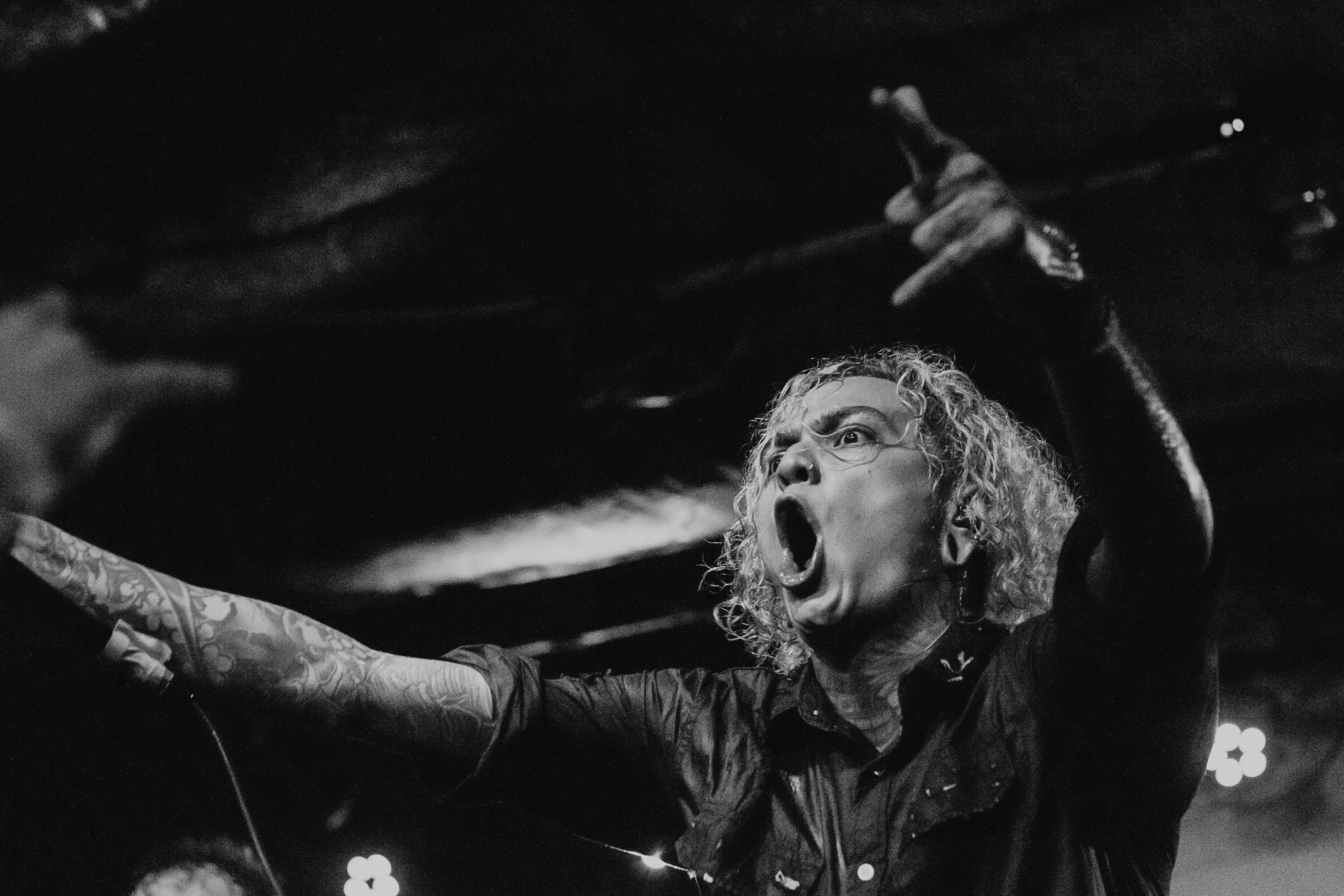 Person with curly hair and glasses shouting with arms raised, in black and white.