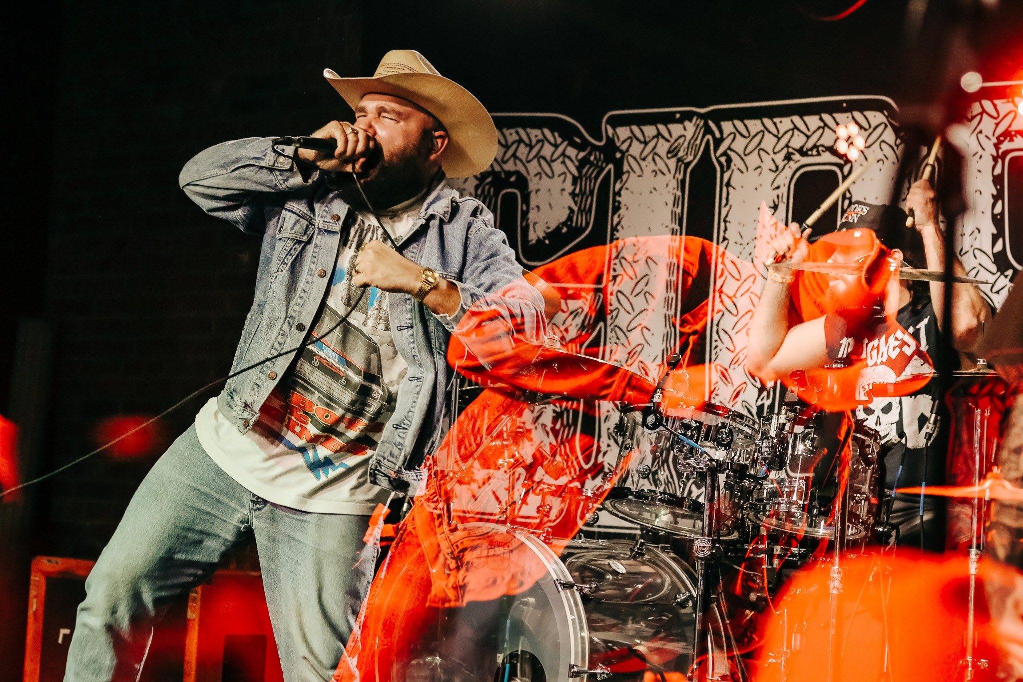 Performer wearing a cowboy hat and denim jacket singing into a microphone on stage with a band behind him.