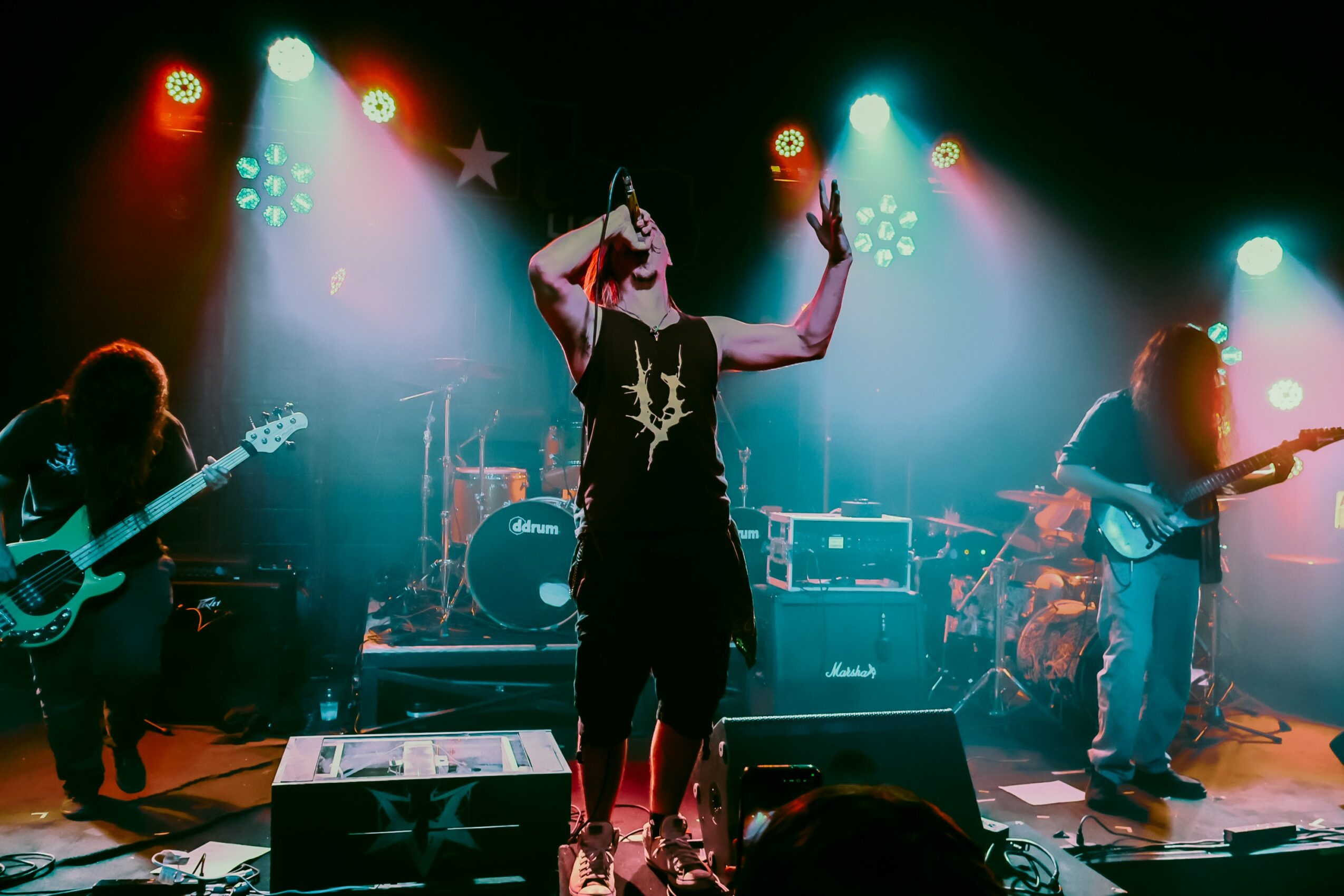 Band performs on stage with colorful lights, singer in center holding microphone, guitarists on sides, drums in background.