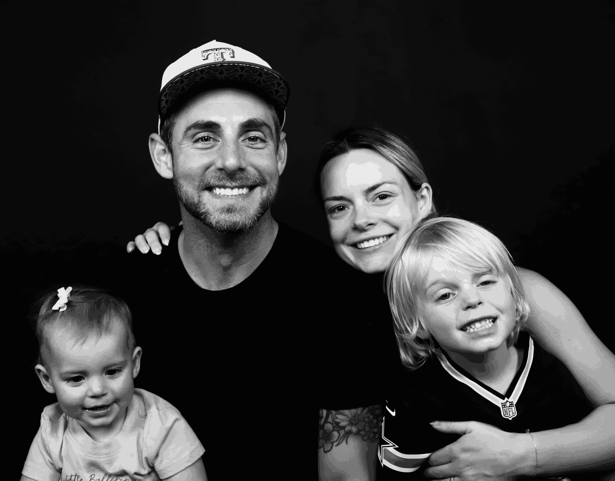 Group of four smiling people, including two children and two adults, against a dark background.