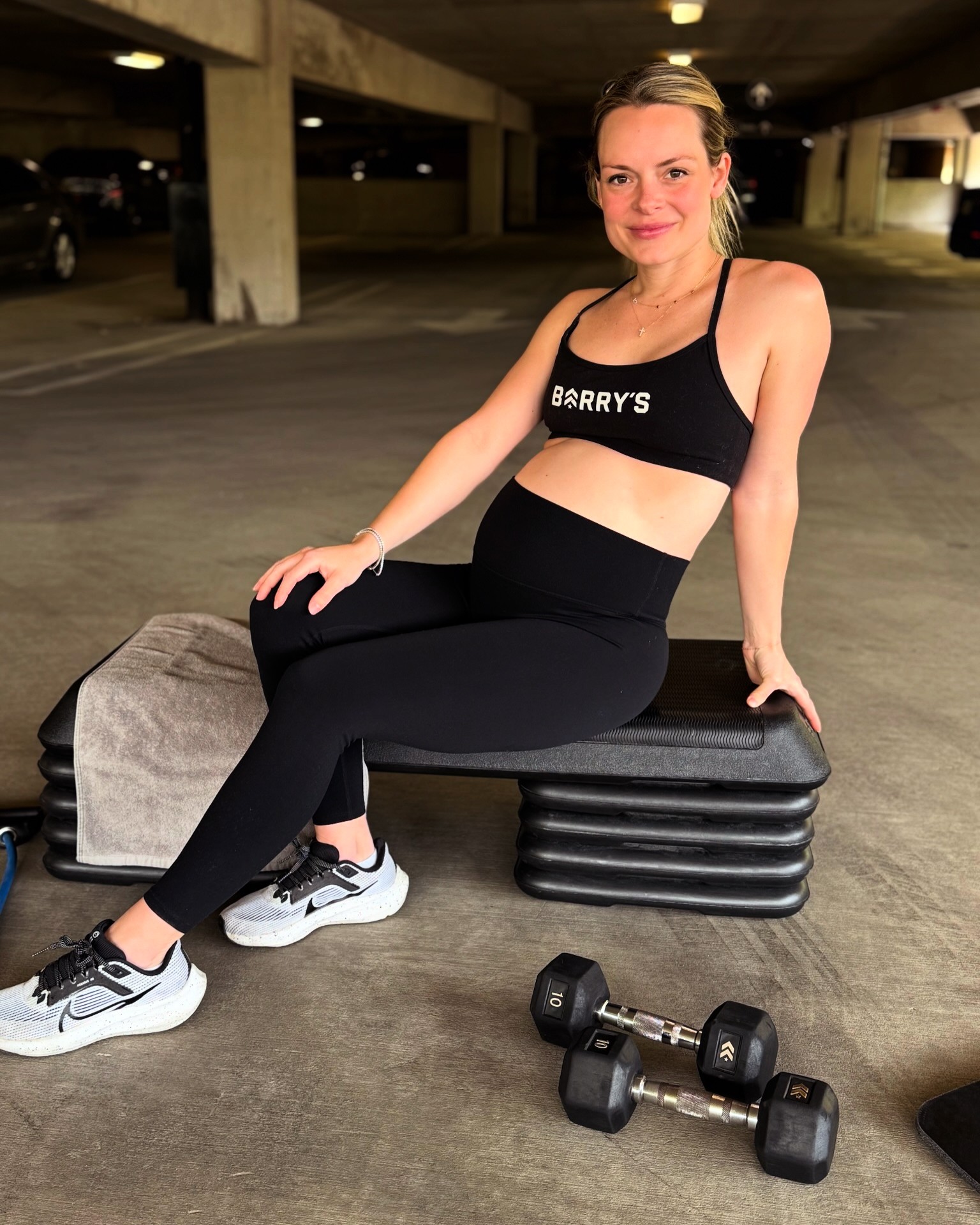 Pregnant woman sitting on a bench in a parking garage, smiling, wearing athletic clothing and sneakers, with dumbbells nearby.