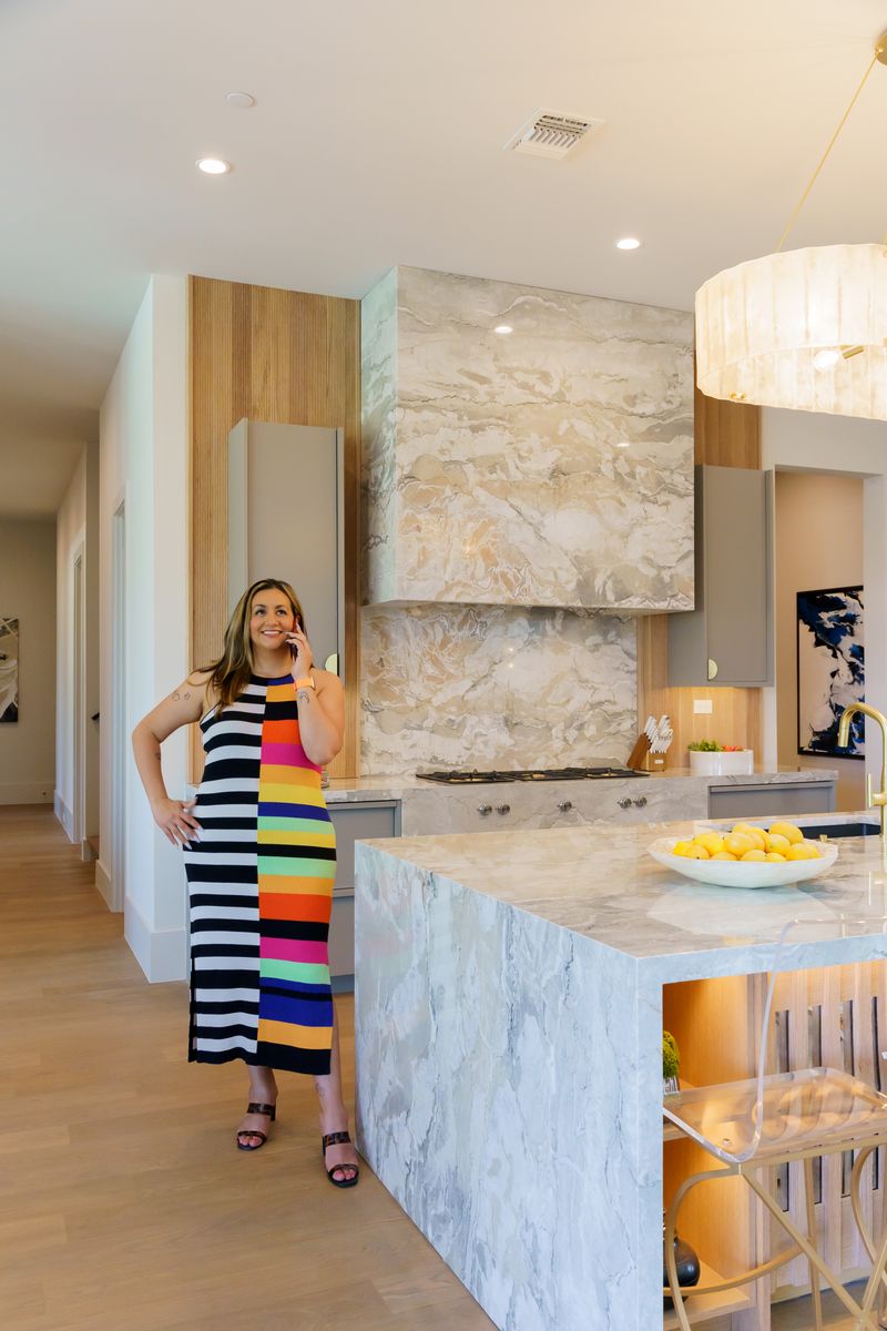 Woman in a colorful striped dress stands in a modern kitchen with marble countertops and a large hanging light fixture.