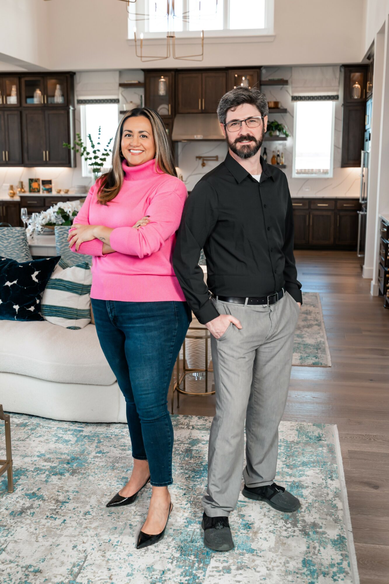 A woman in a pink sweater and jeans, and a man in a black shirt and gray pants, stand in a living room with a kitchen in the background.