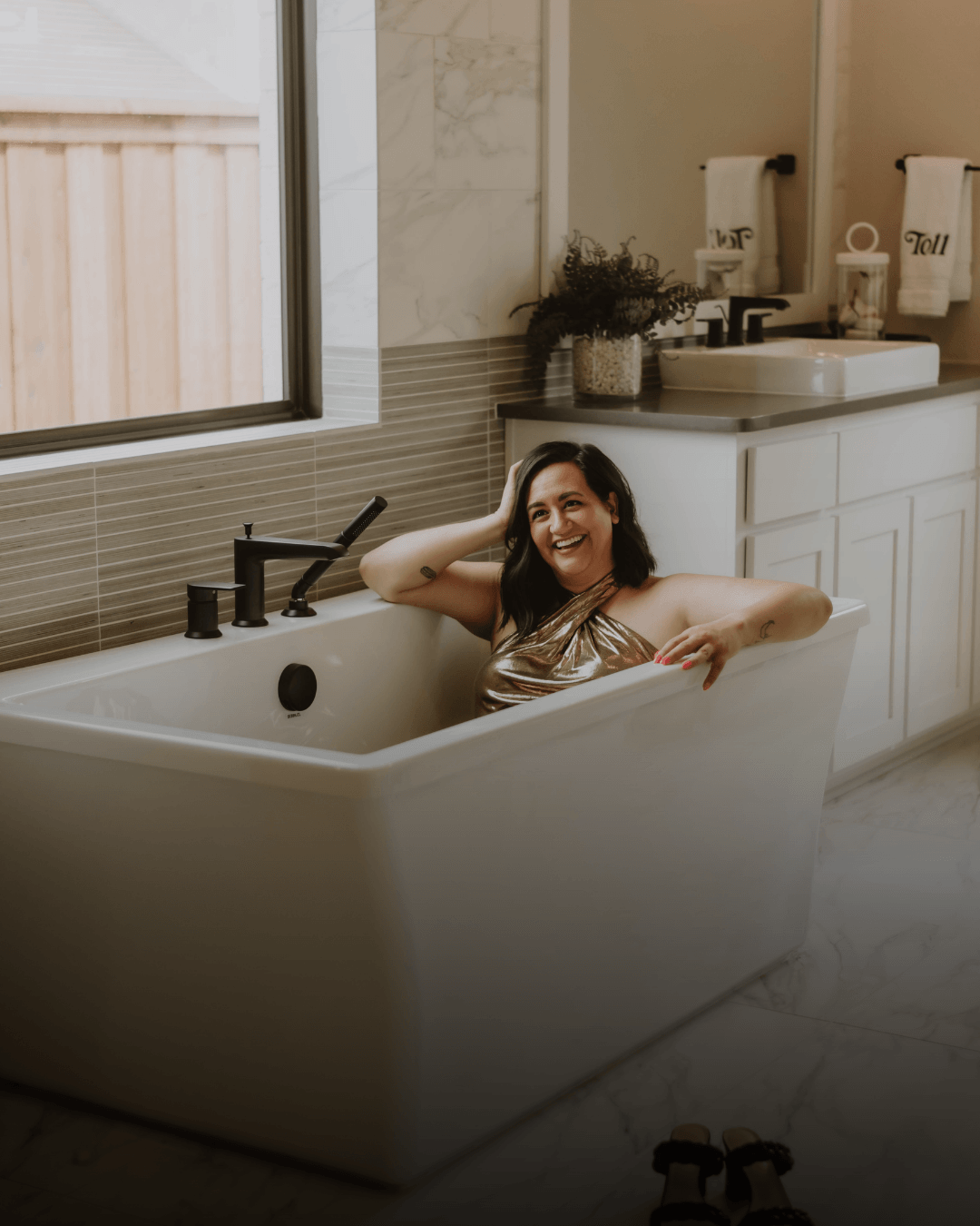 Woman smiling in a bathtub in a bathroom with a window, sink, and plants.