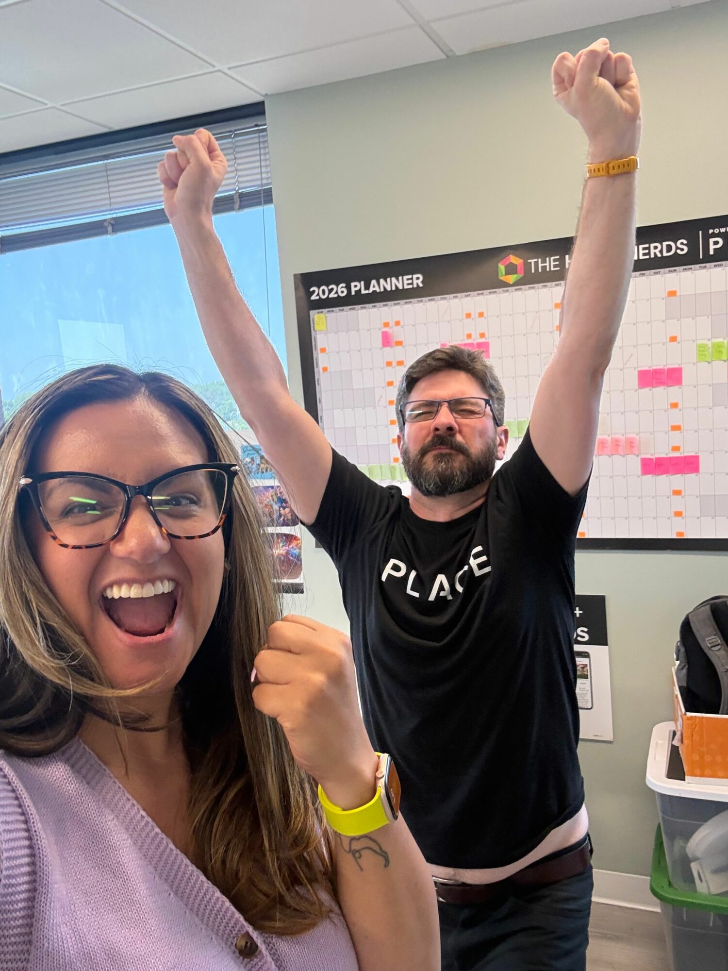 Two people smiling and celebrating indoors, one with glasses and the other with a headband, raising their arms.