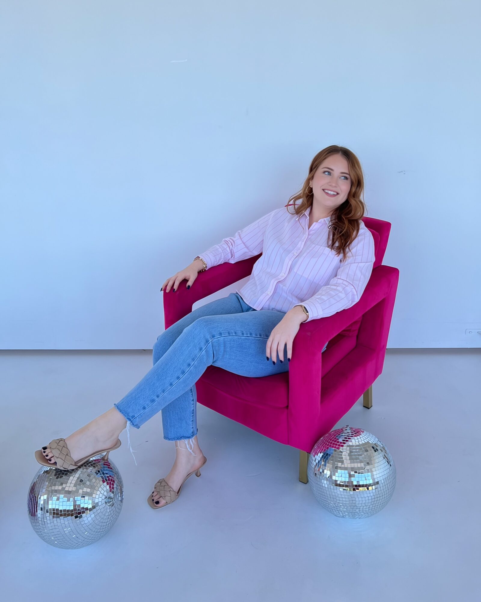 Woman sitting in a pink chair, smiling, with two silver disco balls on the floor, against a plain light blue wall.
