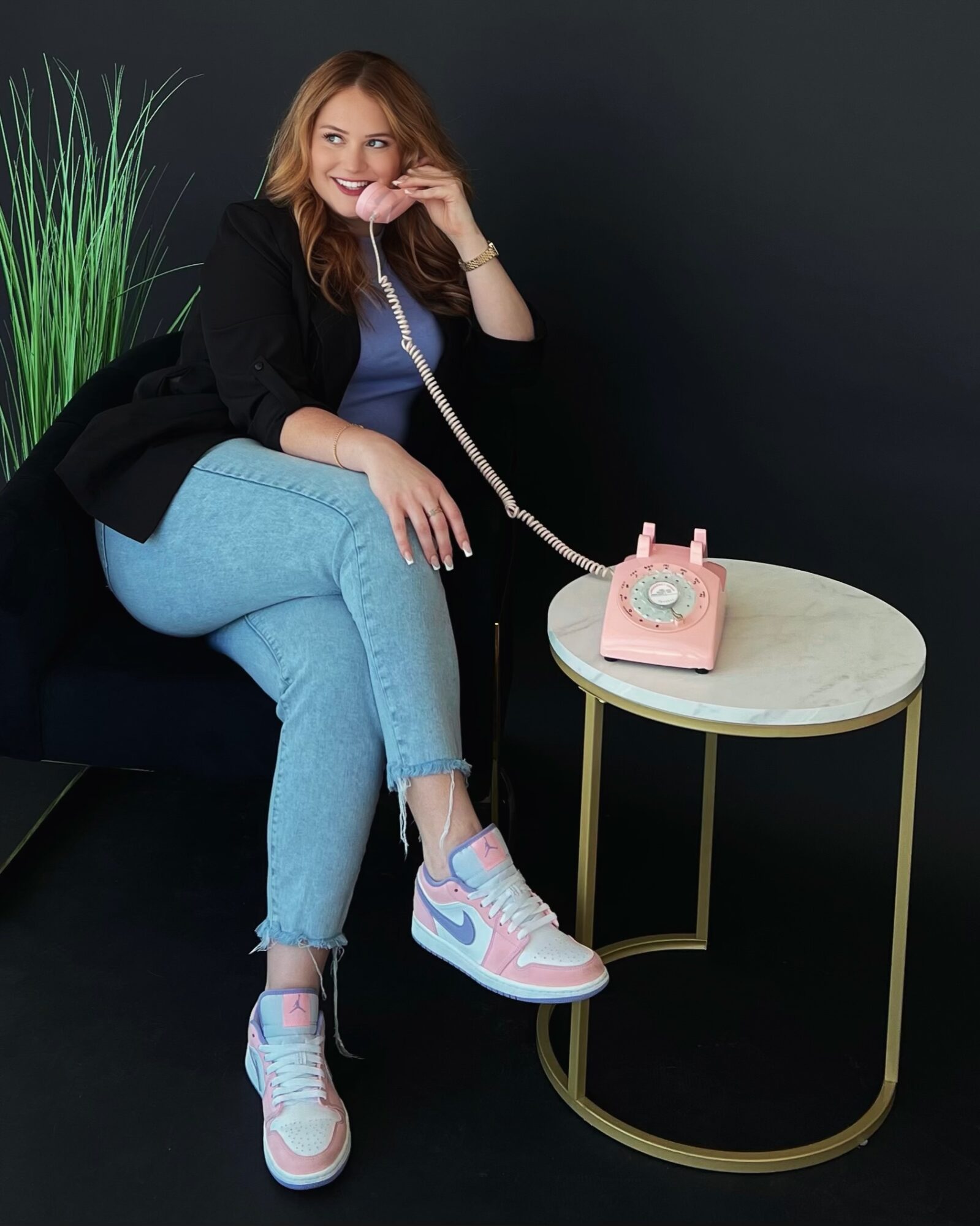 Young woman sitting on a black couch, talking on a pink rotary phone, with a small round table nearby, against a dark background.