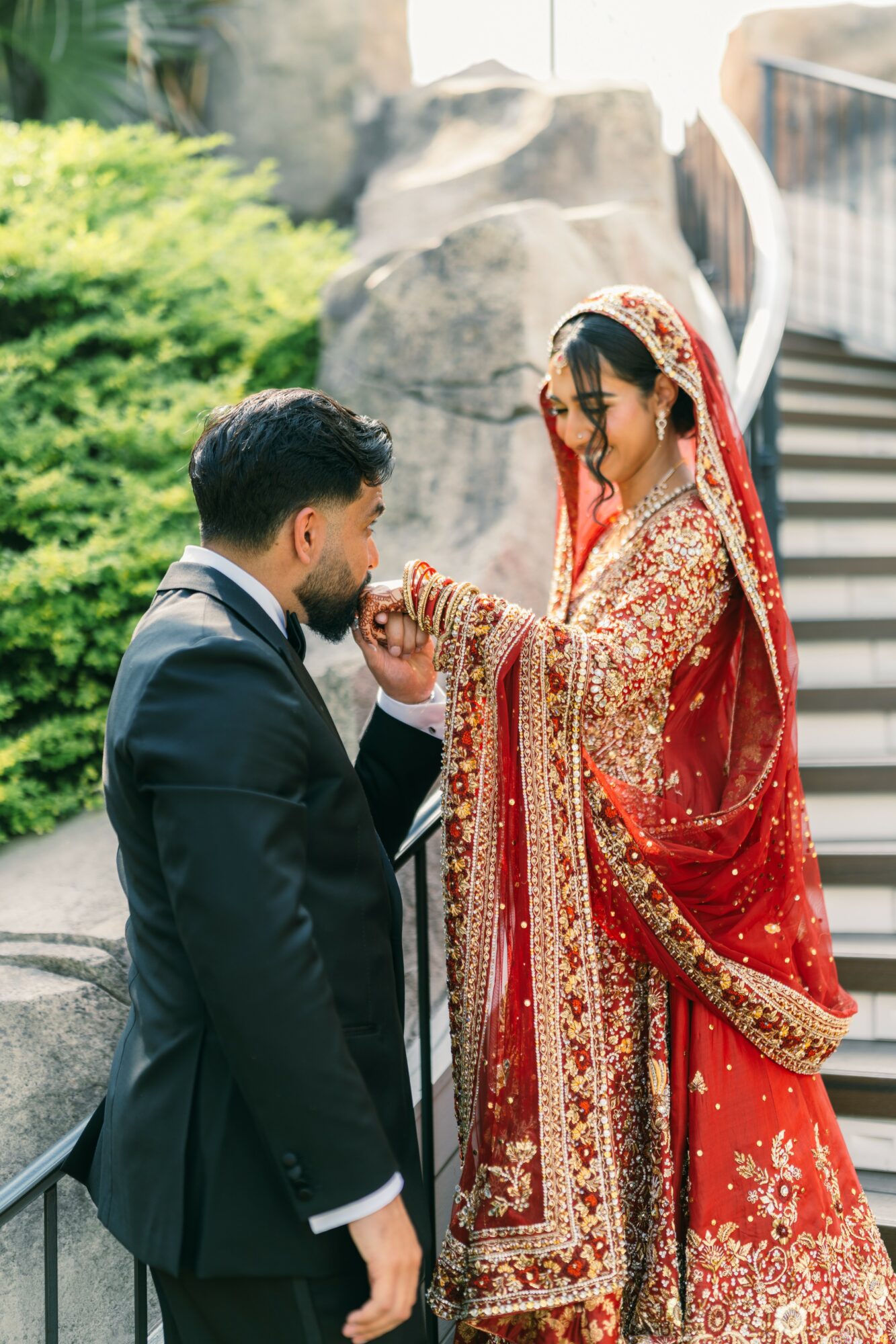 A bride in a red and gold traditional dress and a groom in a black suit are outdoors, with the groom kneeling and the bride holding his hand.