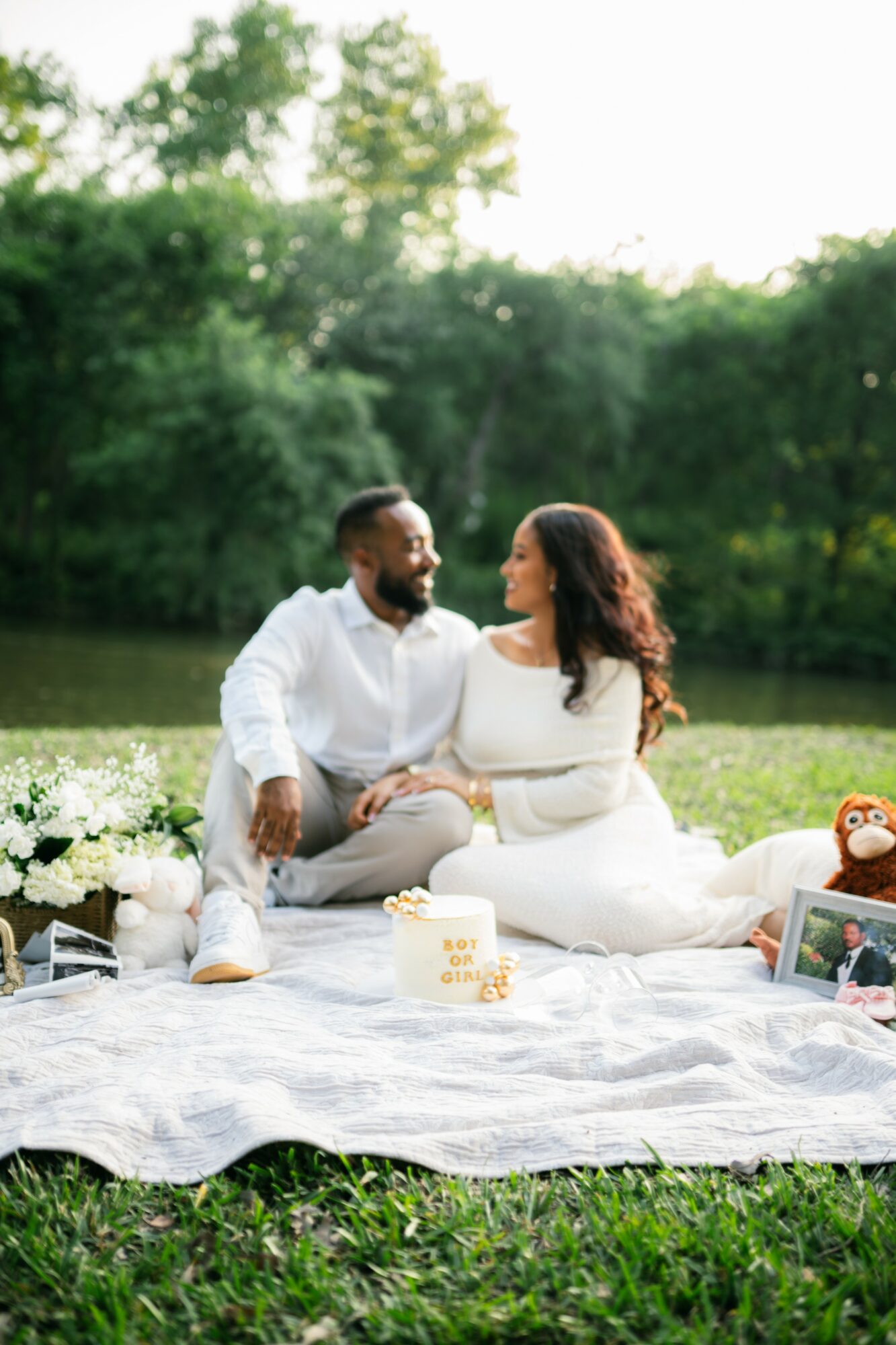 Couple sitting on a blanket outdoors with trees in the background, enjoying a picnic with flowers, a cake, and a framed photo.