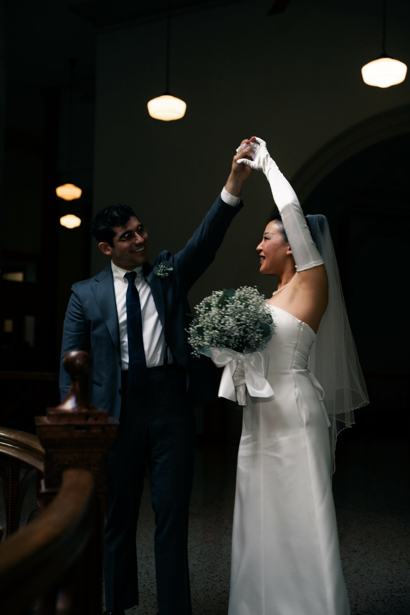 A bride and groom dance indoors, holding hands, with the bride holding a bouquet, under hanging lights.