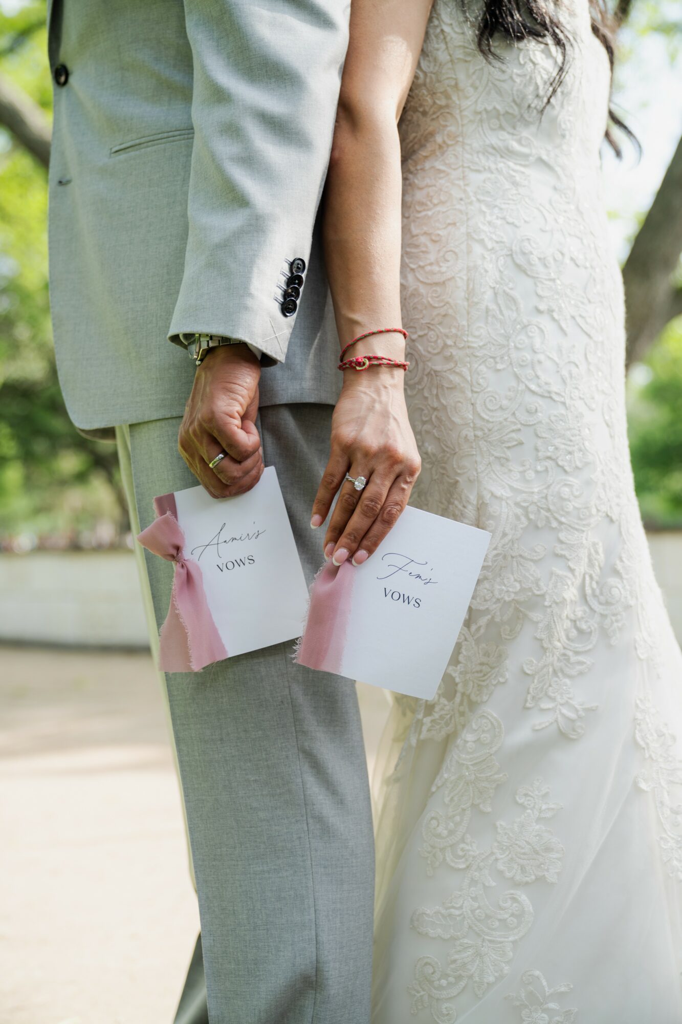 Close-up of a couple holding hands, with wedding attire and holding programs, outdoors with blurred greenery background.