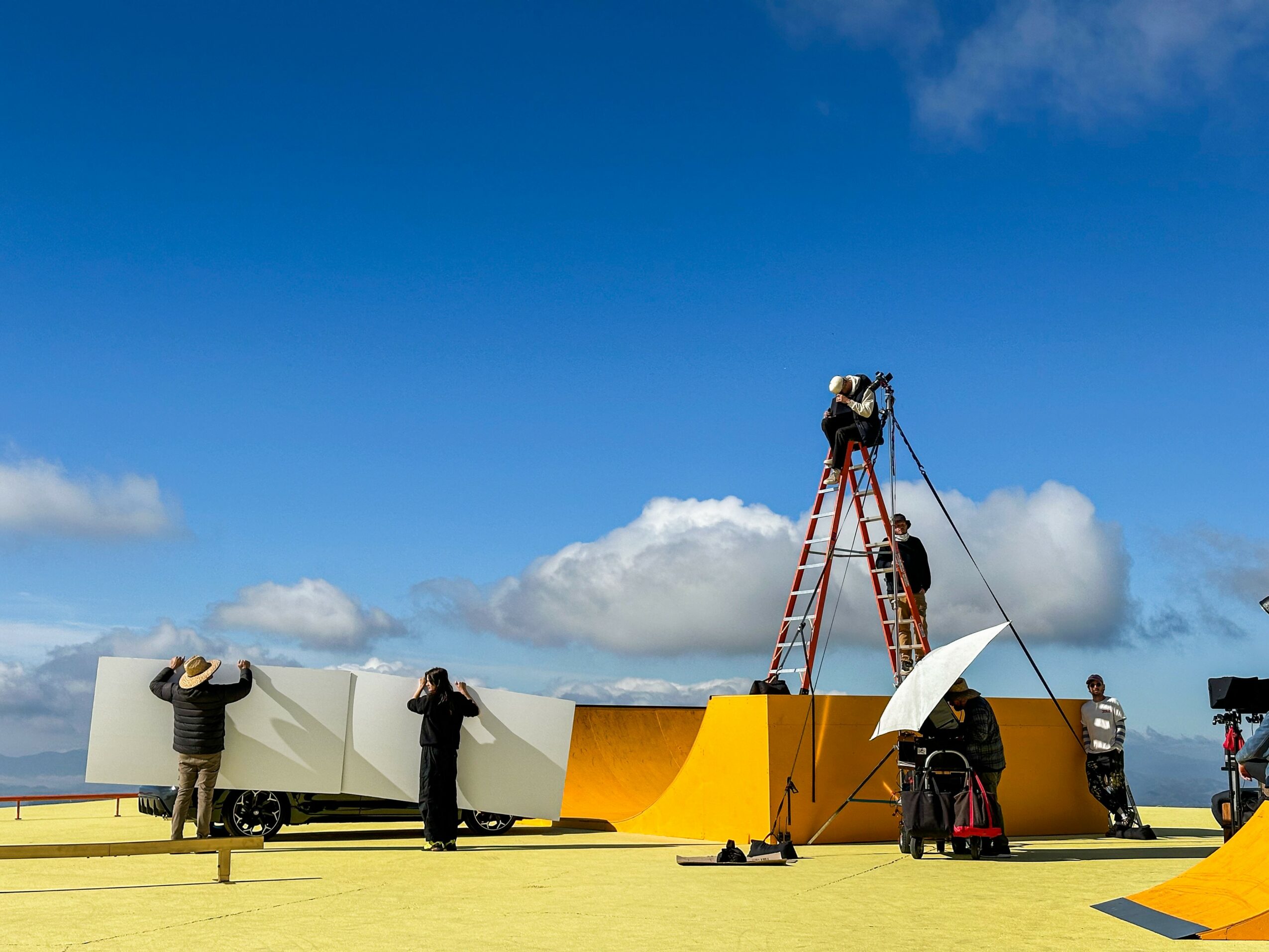 People working on a yellow skate ramp outdoors under a blue sky with clouds, some using tools and equipment.