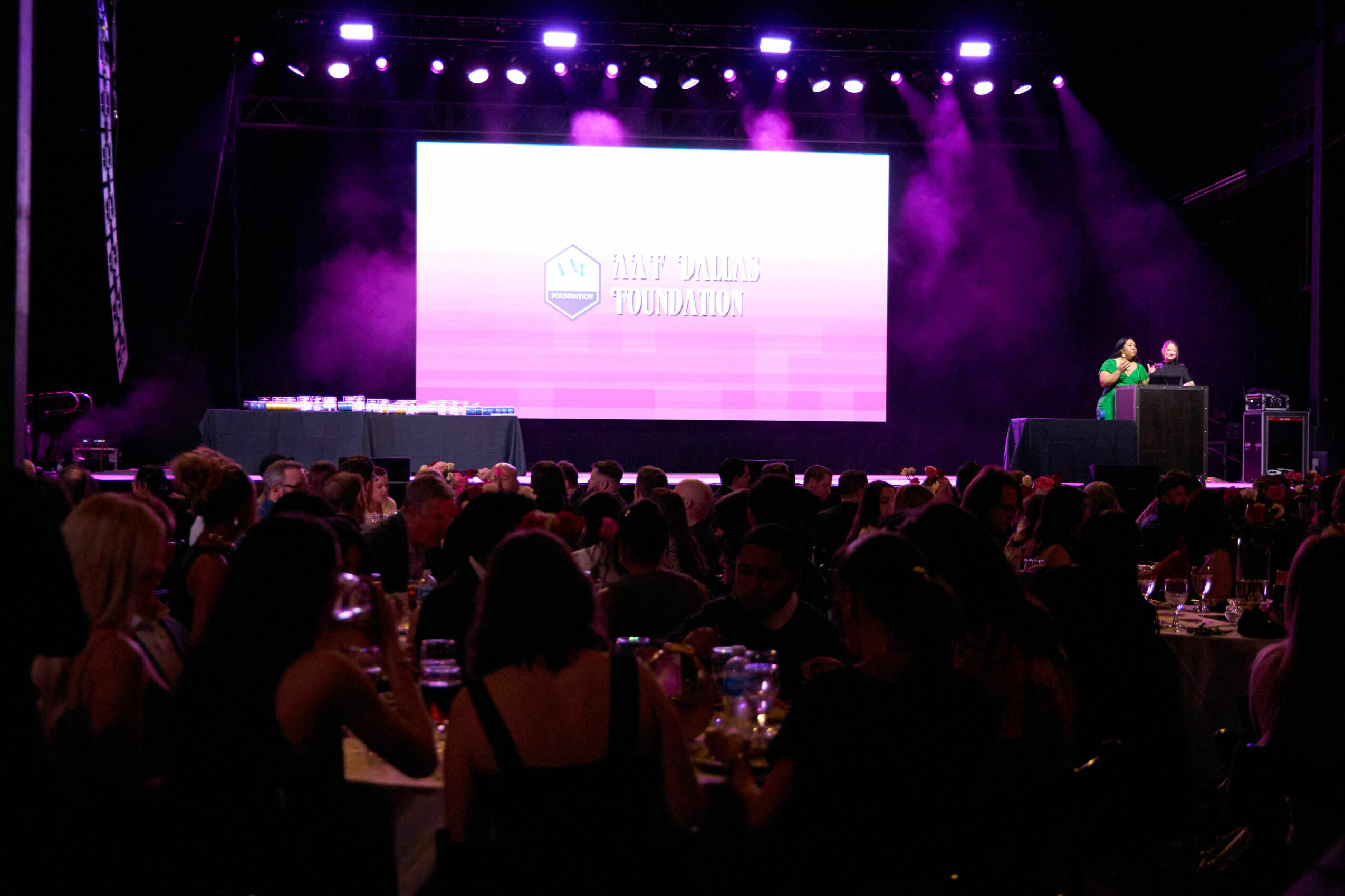 Stage with large screen, purple lighting, and audience seated at tables, event setting.