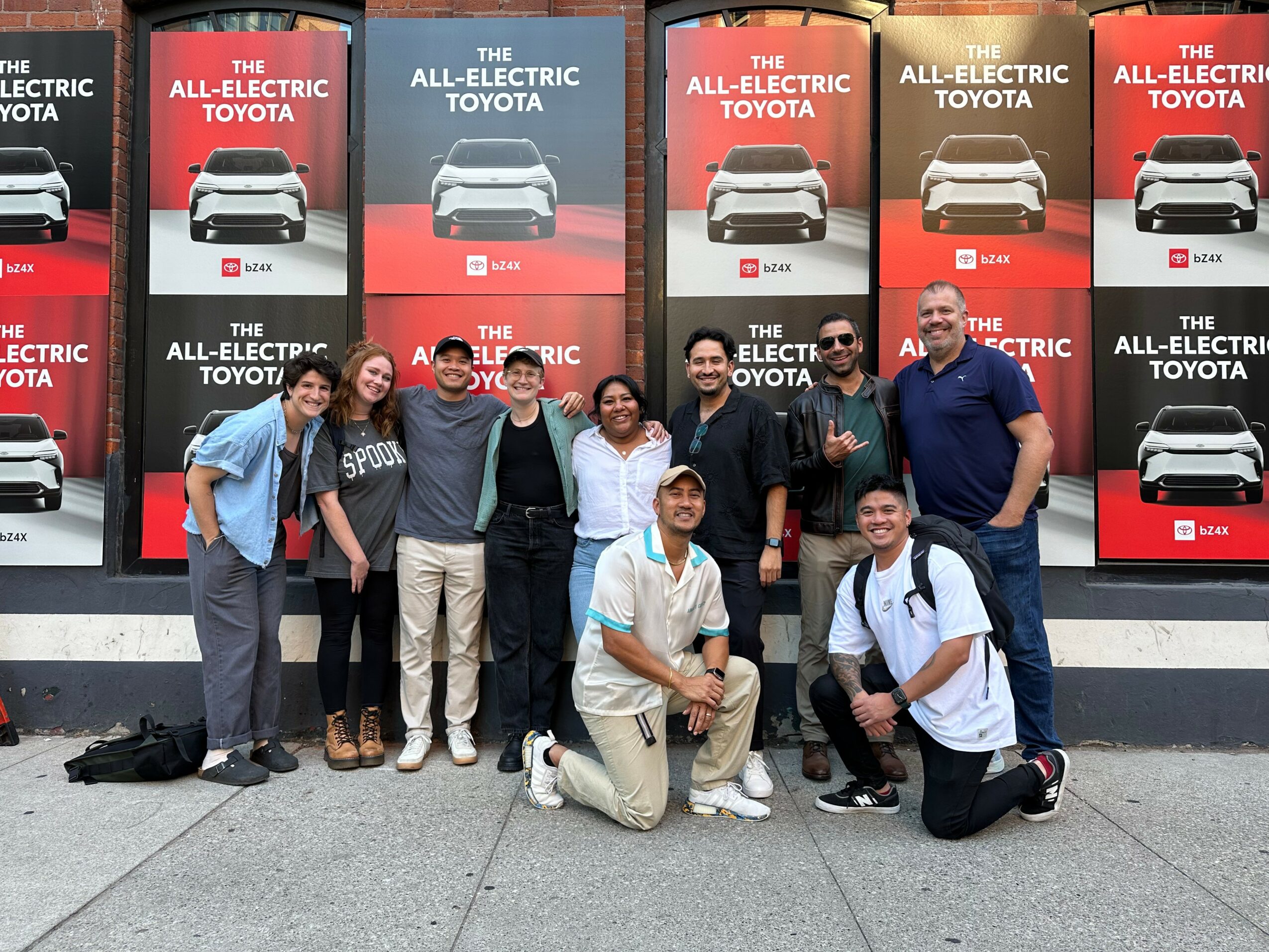 Group of nine people standing and kneeling in front of car advertisements, smiling outdoors.