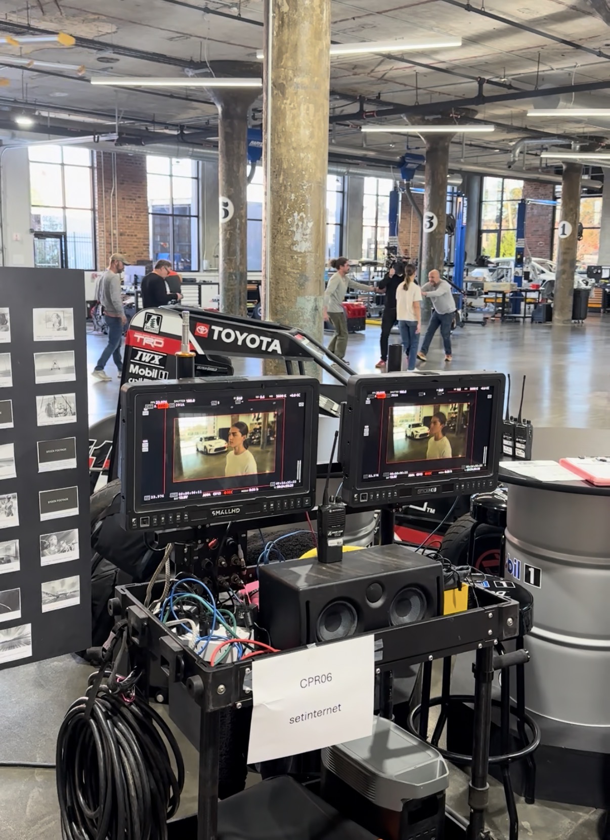 Two monitors on a cart display a person in a room with large windows and industrial interior, with people and equipment in background.