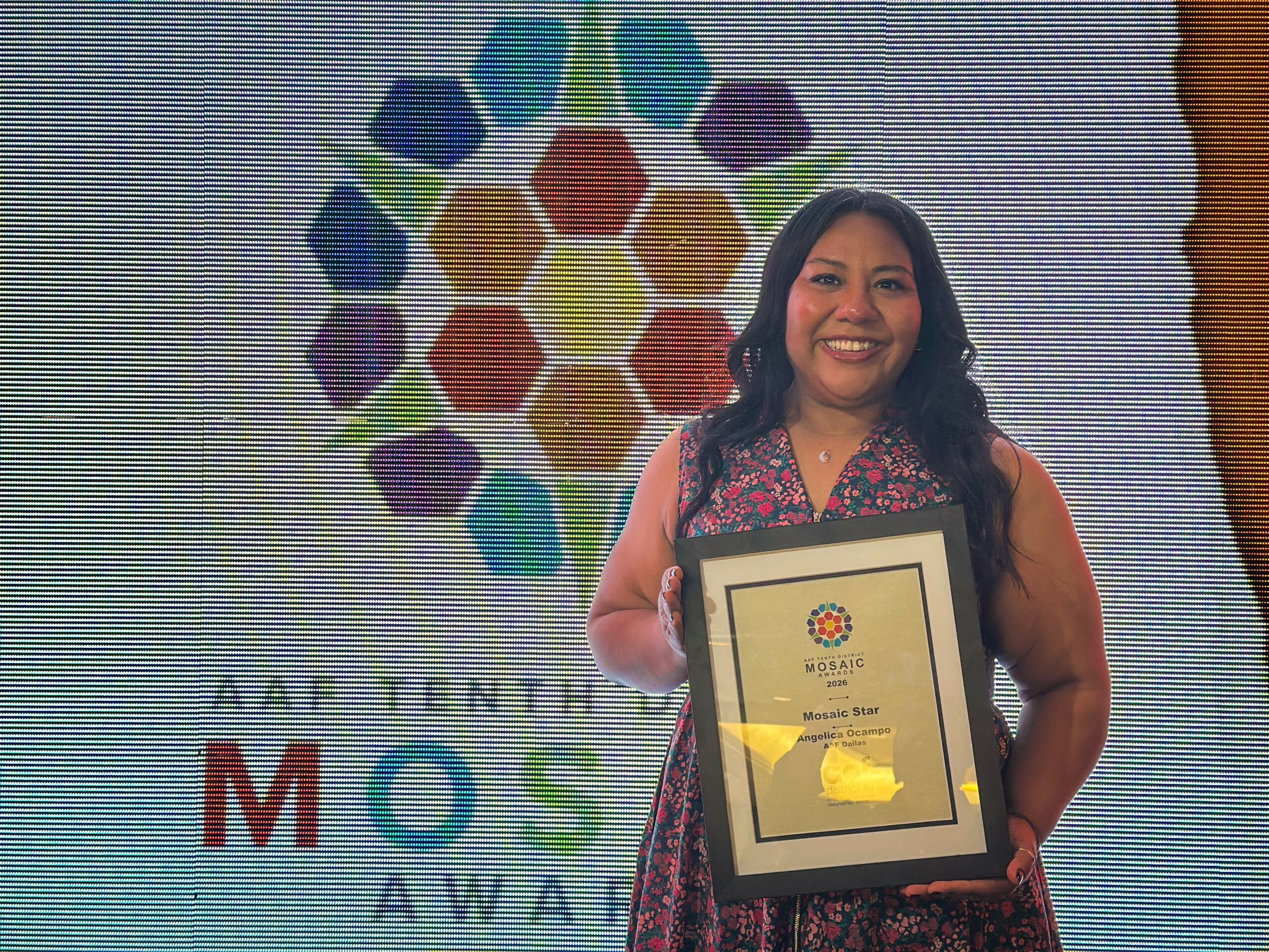 Woman holding an award plaque, smiling, standing in front of a colorful logo and text background.